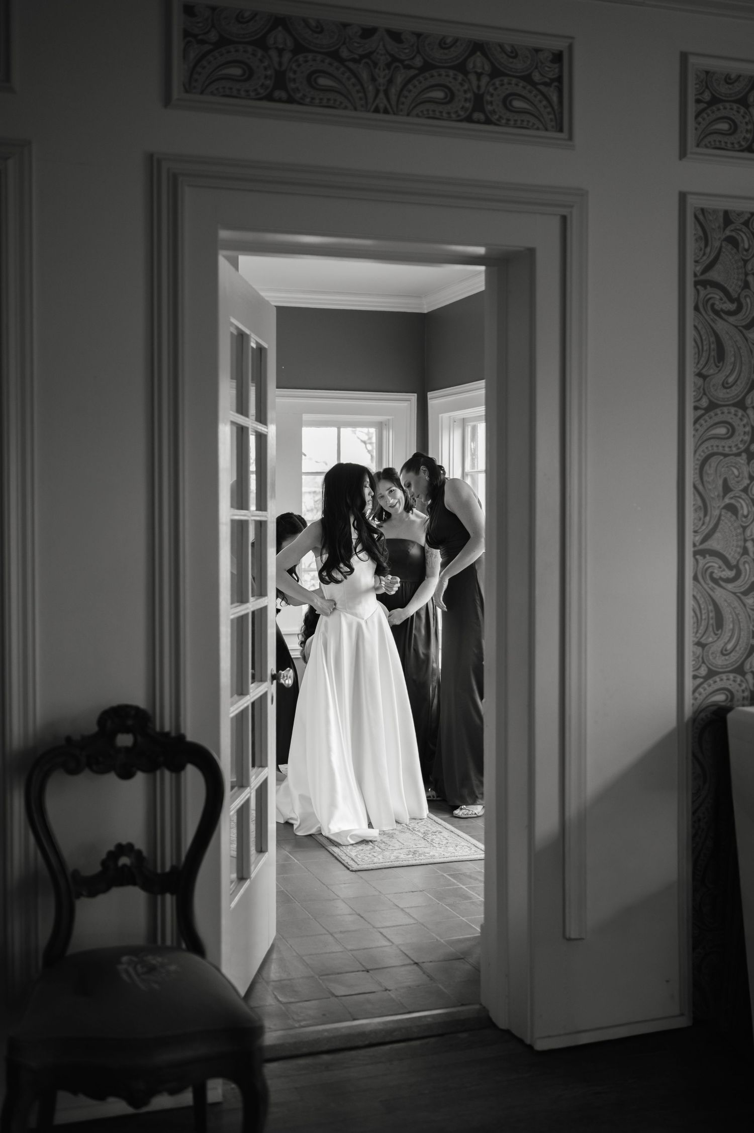 Bride in white gown gets assisted by bridesmaids through elegant doorway in black and white photo.
