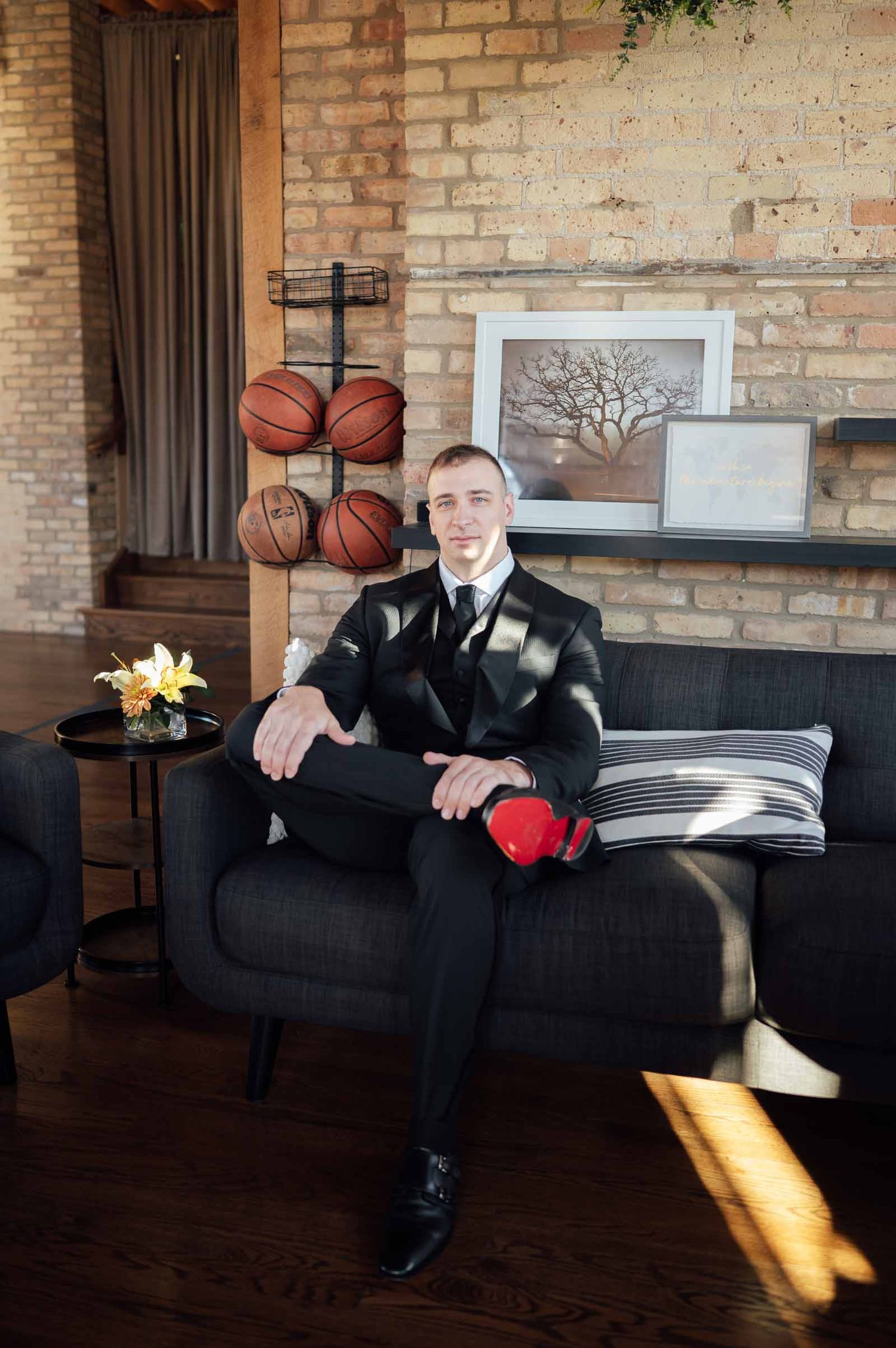 Groom in black suit sits on a sofa with boxing gloves beside a basketball display wall.