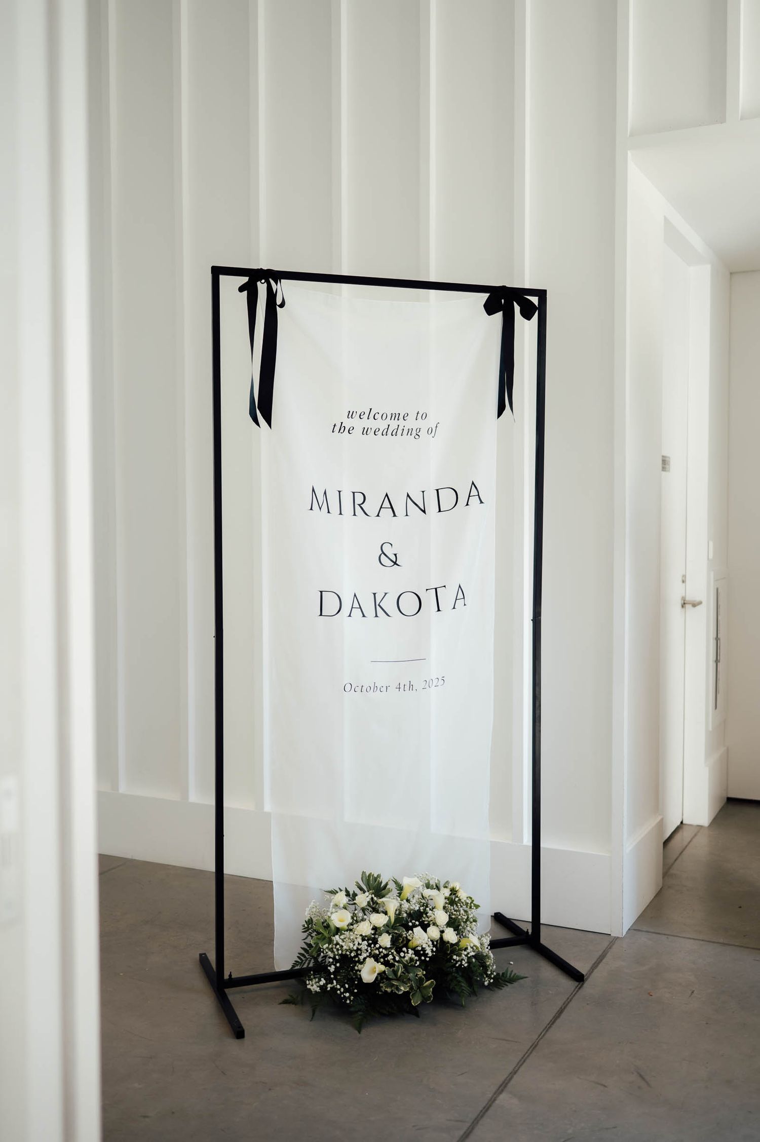 Elegant black and white wedding welcome sign for Miranda and Dakota on a minimalist metal stand with floral arrangement.