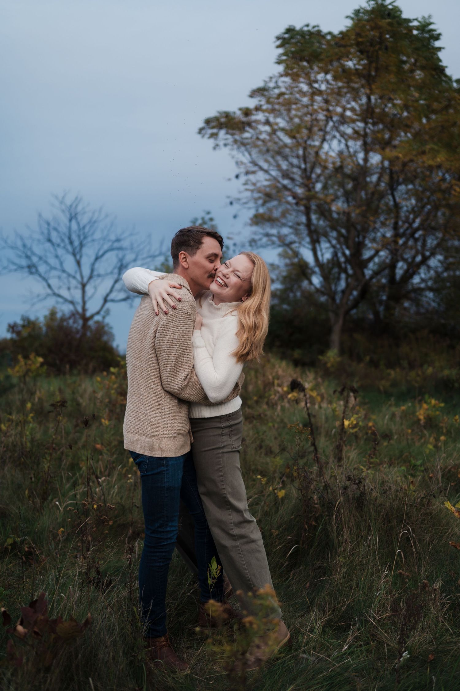 Happy couple embracing and laughing together in an autumn field during a romantic engagement session.