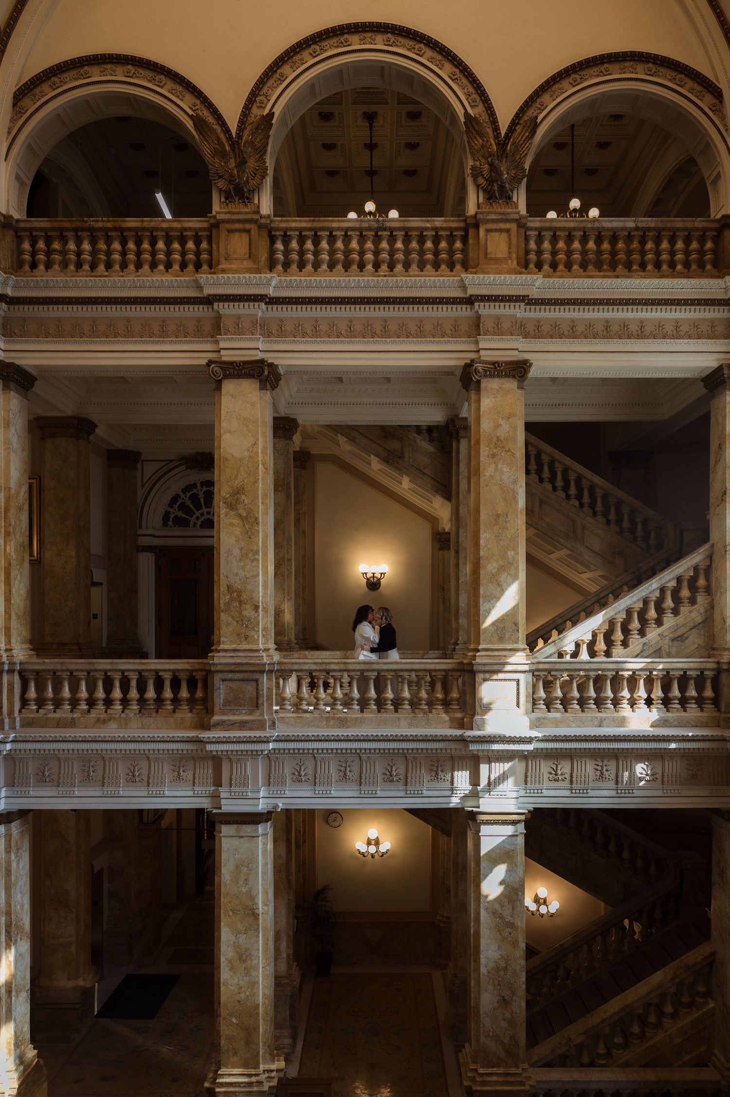 Couple embracing on upper balcony of ornate classical building with arched marble columns.