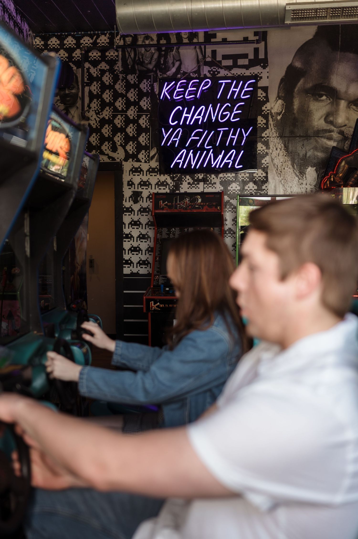 Couple playing vintage arcade games side by side beneath a neon sign reading Keep the Change Ya Filthy Animal.