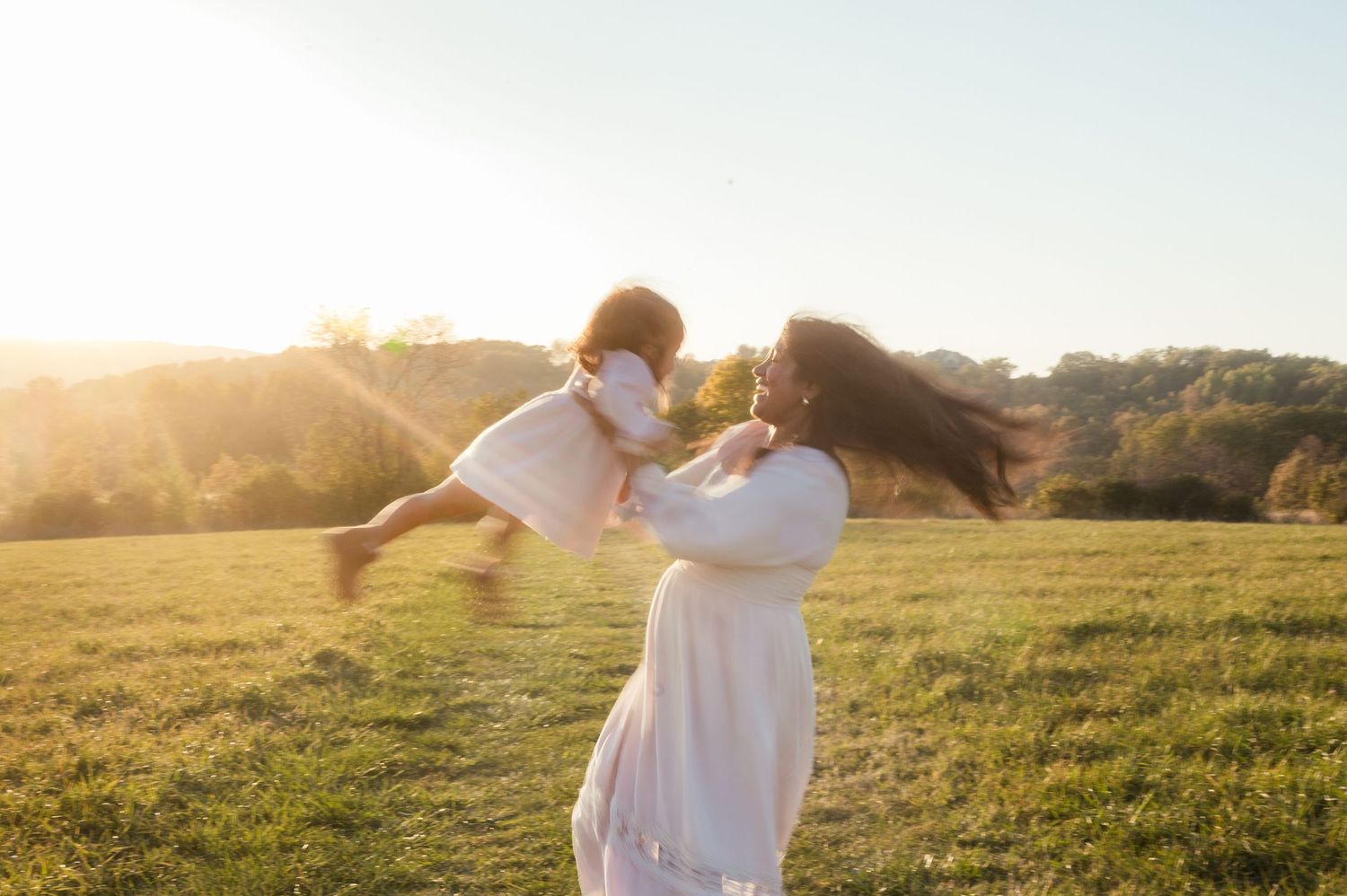 Mother lifting happy toddler in golden sunset light in a green field wearing white dresses.