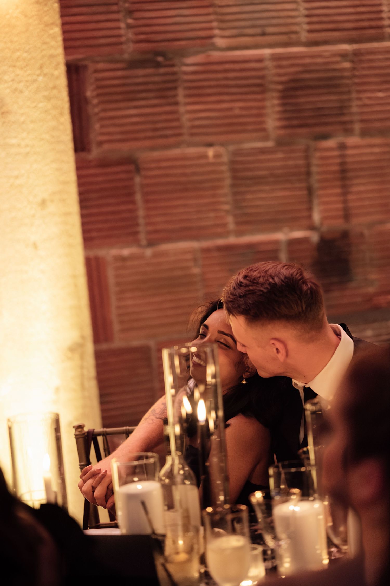 Couple sharing a romantic moment at a candlelit wedding reception table against a warm brick wall backdrop.