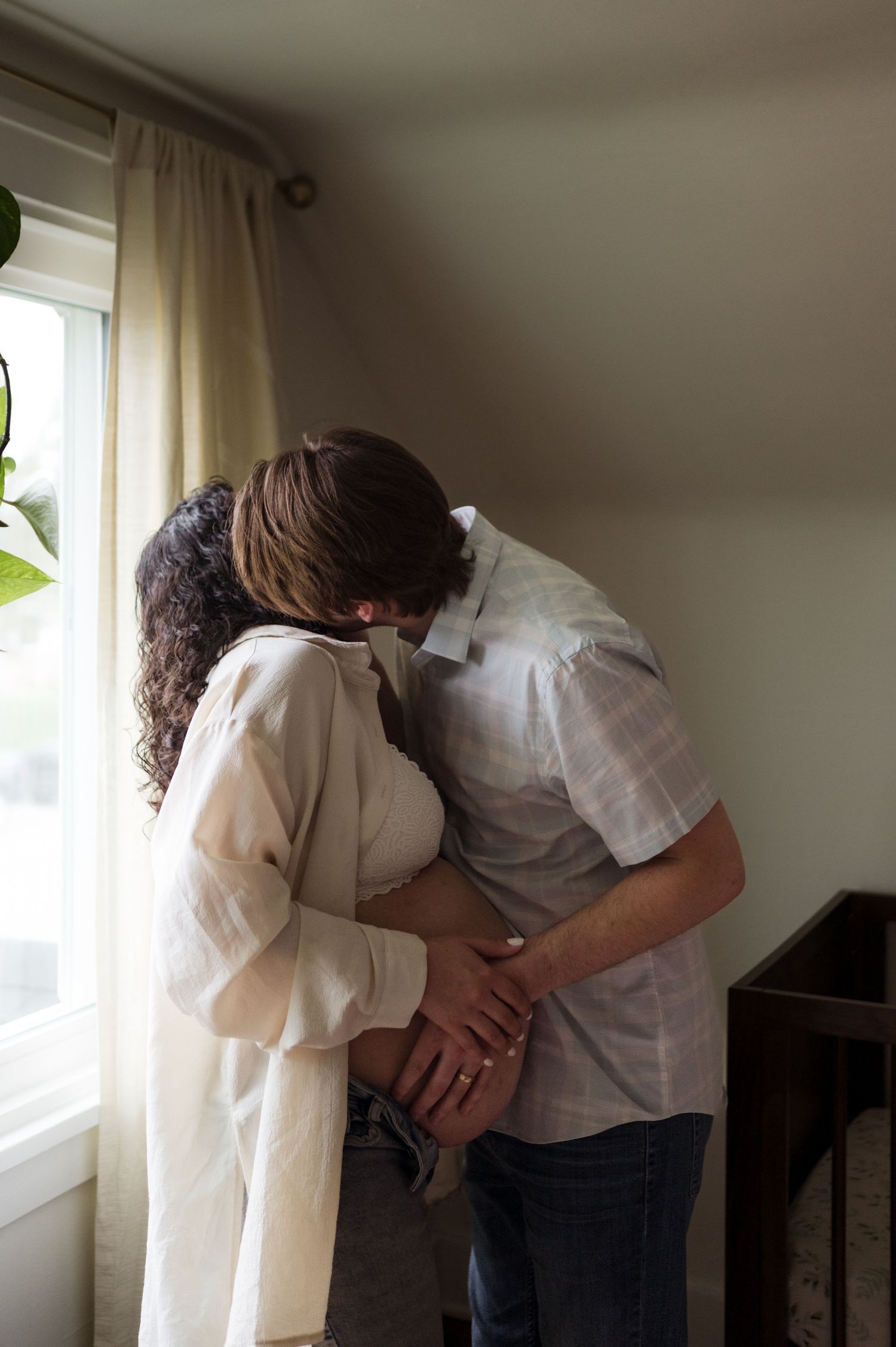 Two people in white shirts share an intimate moment by a window with curtains in a dimly lit room.