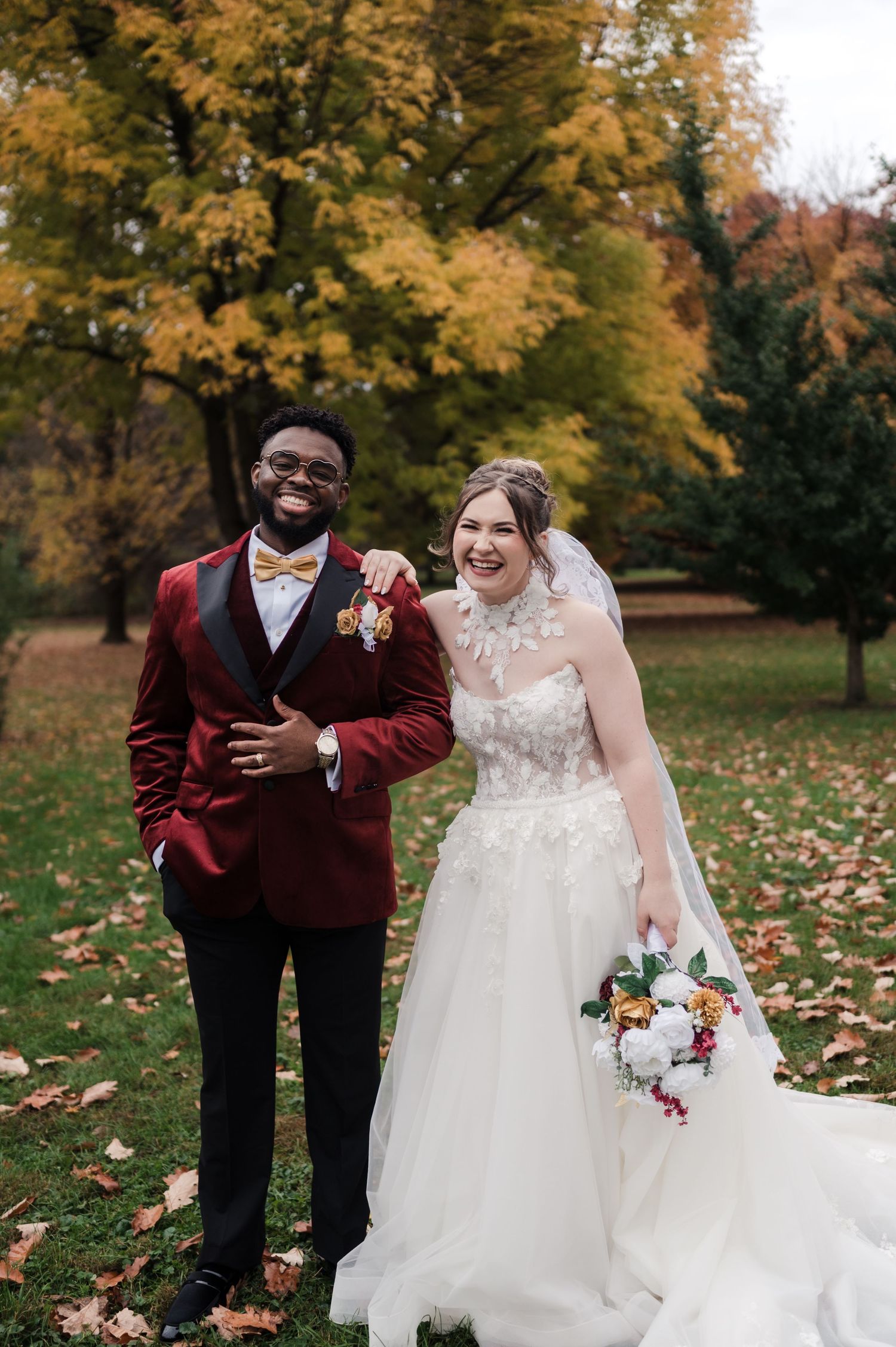 A couple poses together in an autumn park with golden leaves and trees, wearing burgundy and white wedding attire.