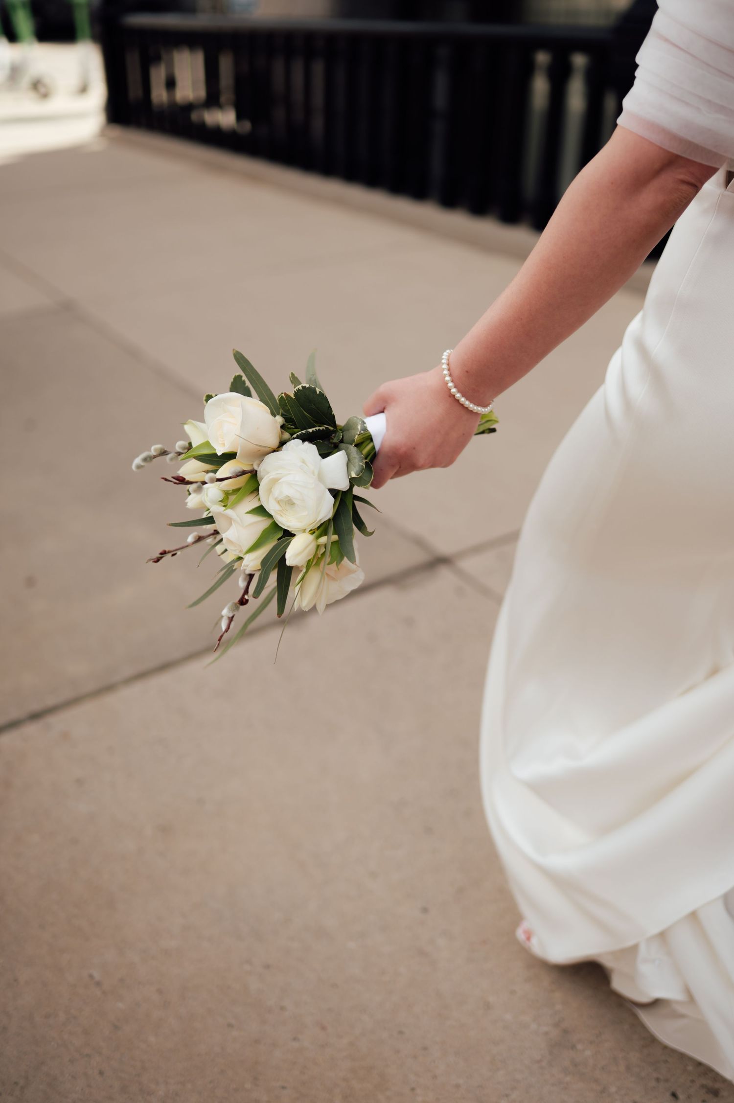 Bride in white gown holding a small bouquet of white roses and greenery while walking on a stone pathway.