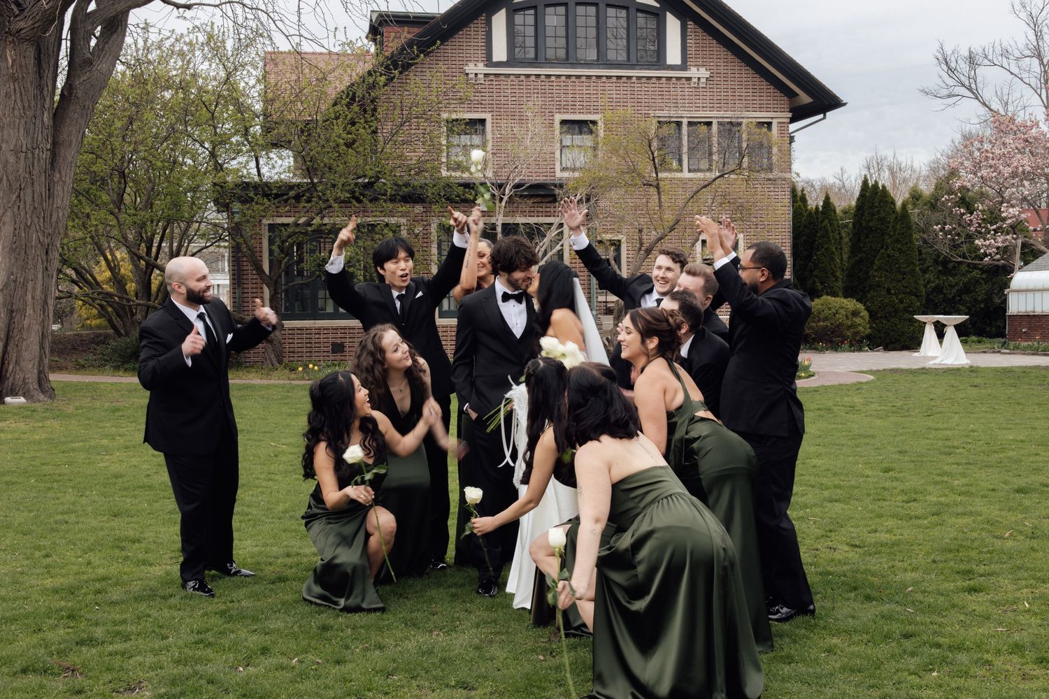 Group of teens in formal prom attire celebrating outdoors on grass in front of a brick house in spring.