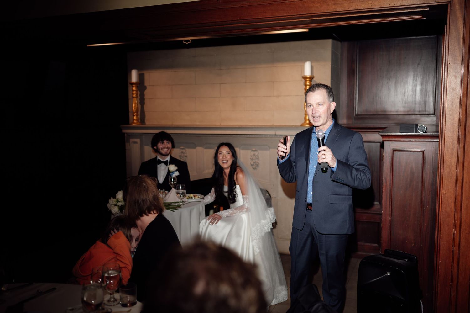 Man giving a toast speech at a wedding reception with bride and groom seated at the head table.