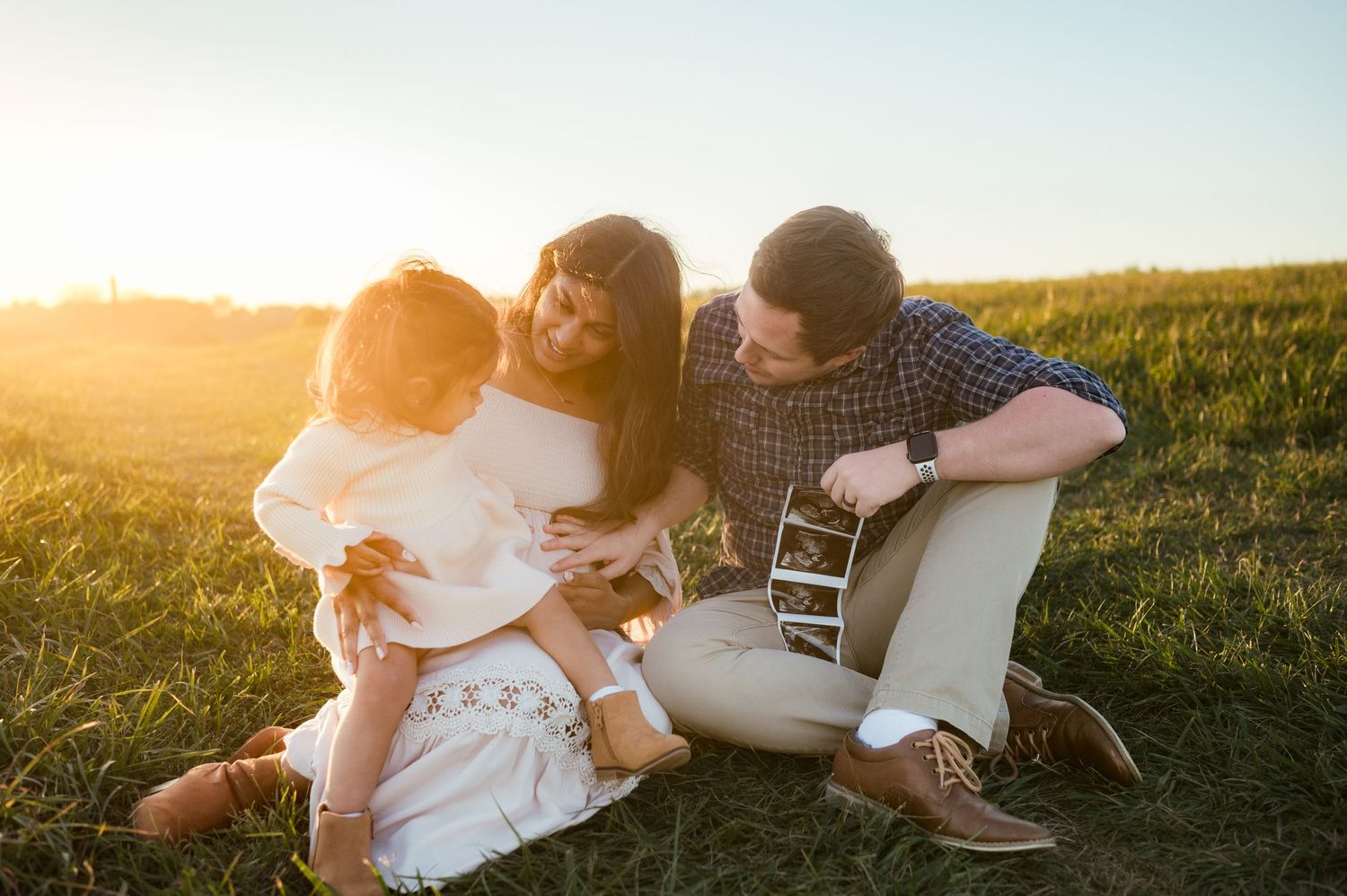 Happy family sitting in sunlit field holding ultrasound photos during pregnancy announcement session.