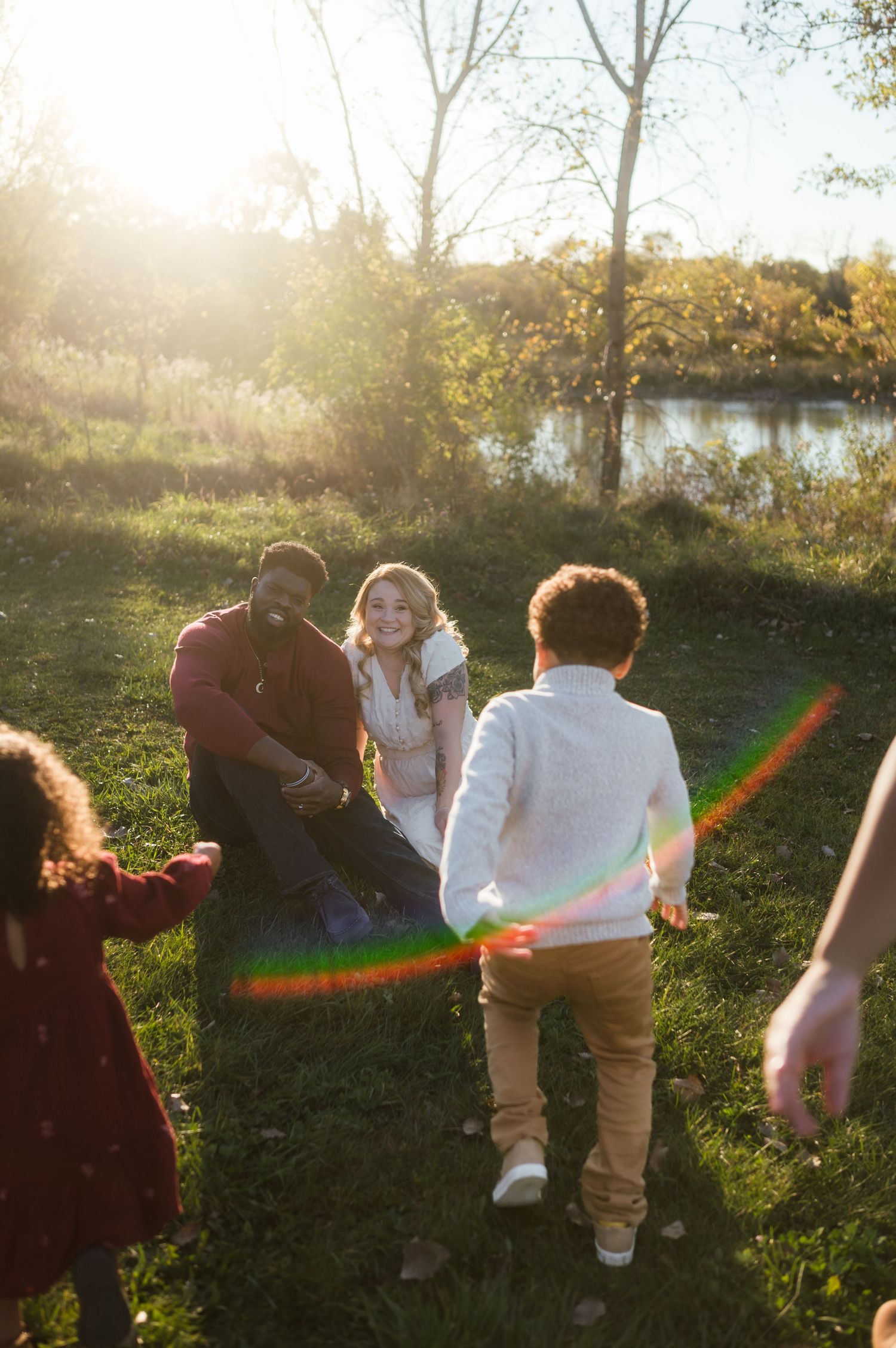 Parents sitting on grass smiling as children run toward them near sunlit pond in autumn.