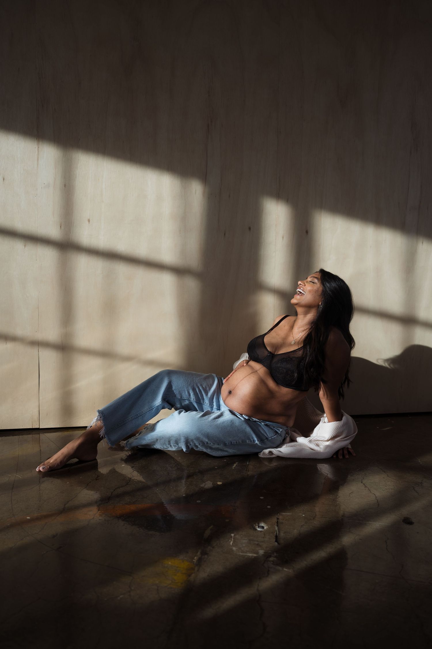 Pregnant woman in black bralette and blue jeans sits on floor laughing in sunlight.