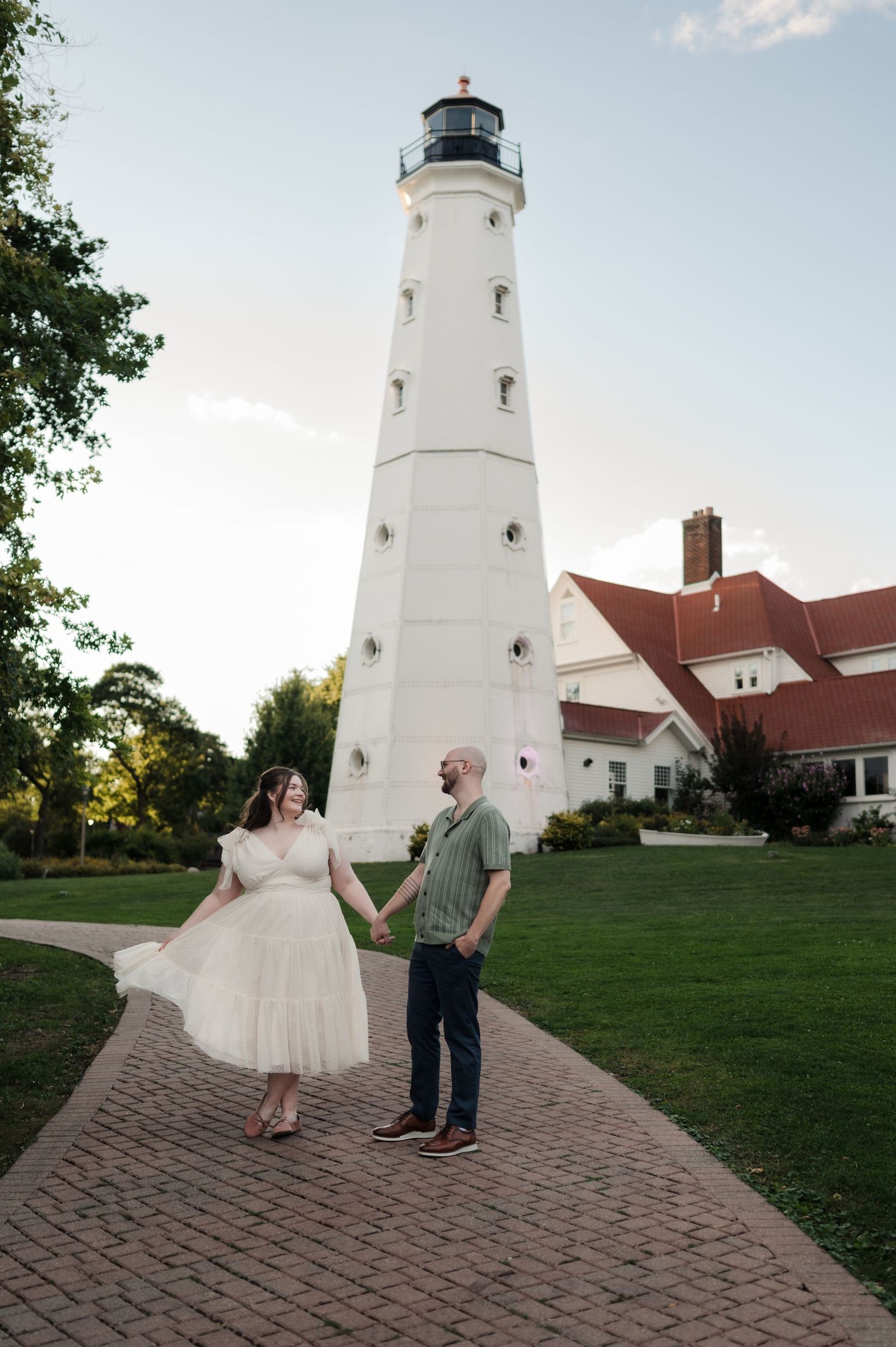 Engaged couple holding hands and walking on brick path in front of tall white lighthouse at sunset.