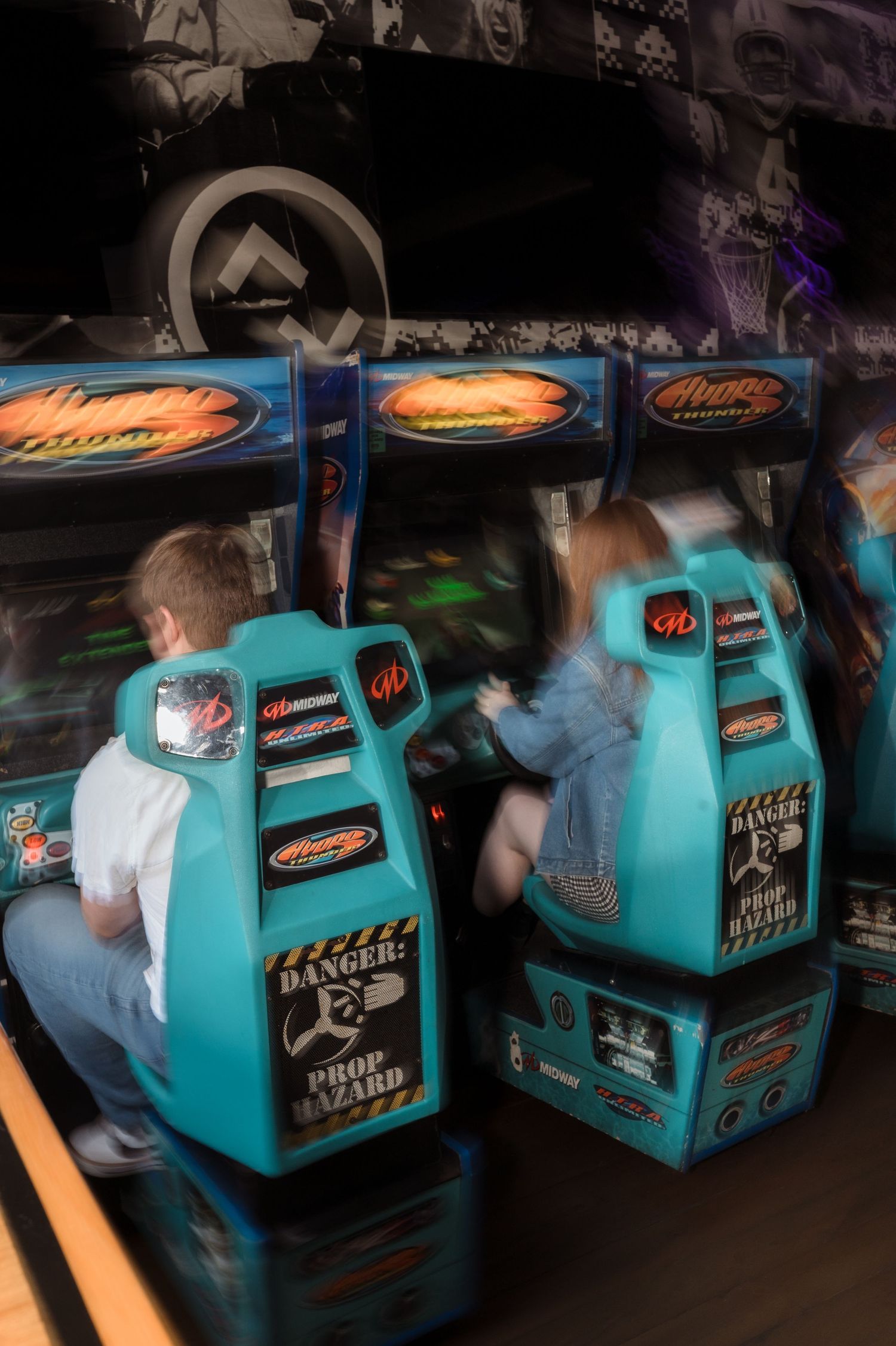 Two children playing teal arcade racing games with Prop Hazard warning labels in a dark arcade.