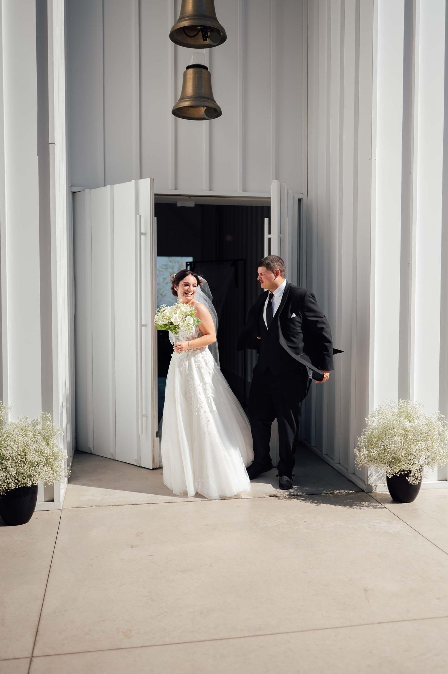 Smiling bride and groom exit through grand white chapel doors flanked by baby's breath floral arrangements.