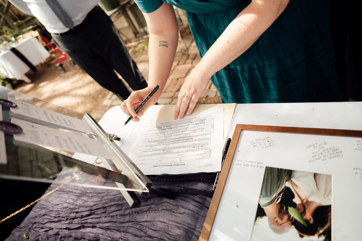 Person in teal dress signing wedding documents at an outdoor ceremony table with photos displayed nearby.