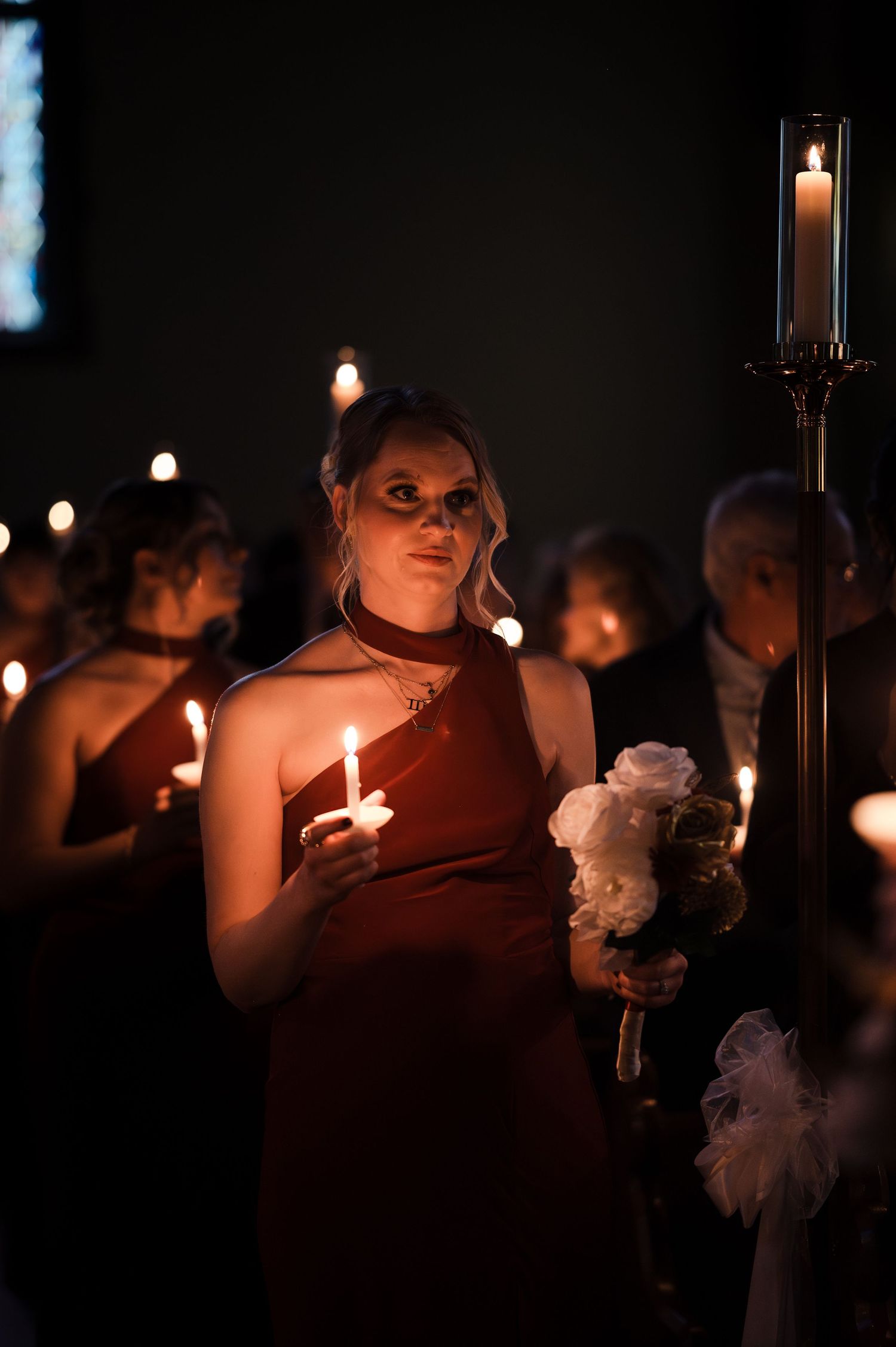 Bridesmaid in red dress holding candle and white flowers during candlelit wedding ceremony.