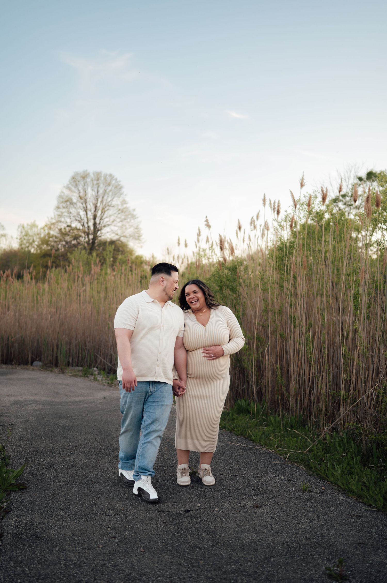 Expecting couple laughing and walking hand in hand on a path beside tall reeds at dusk.