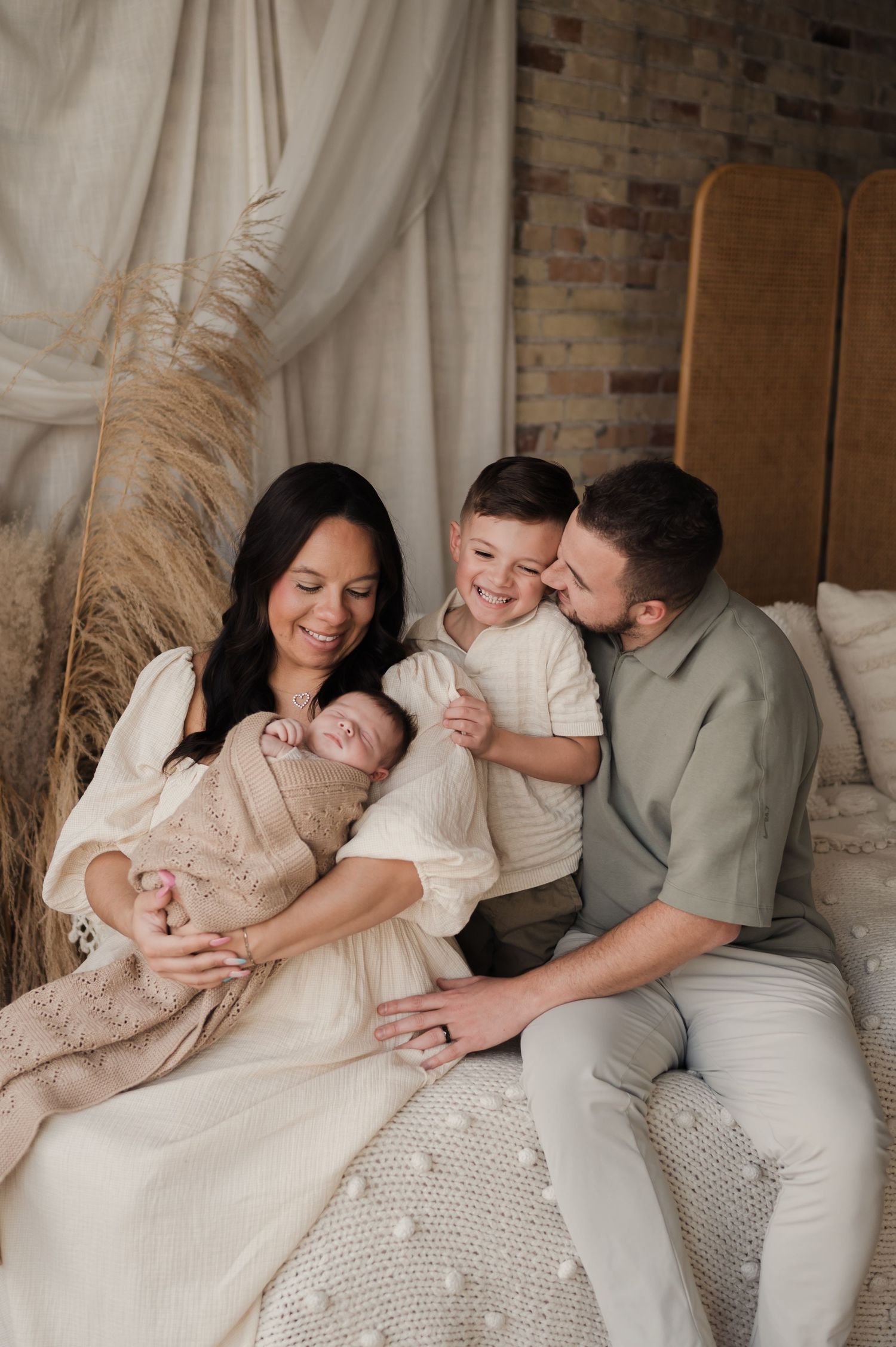 A happy family of four sits together on a bed, parents smiling as their toddler hugs the newborn.
