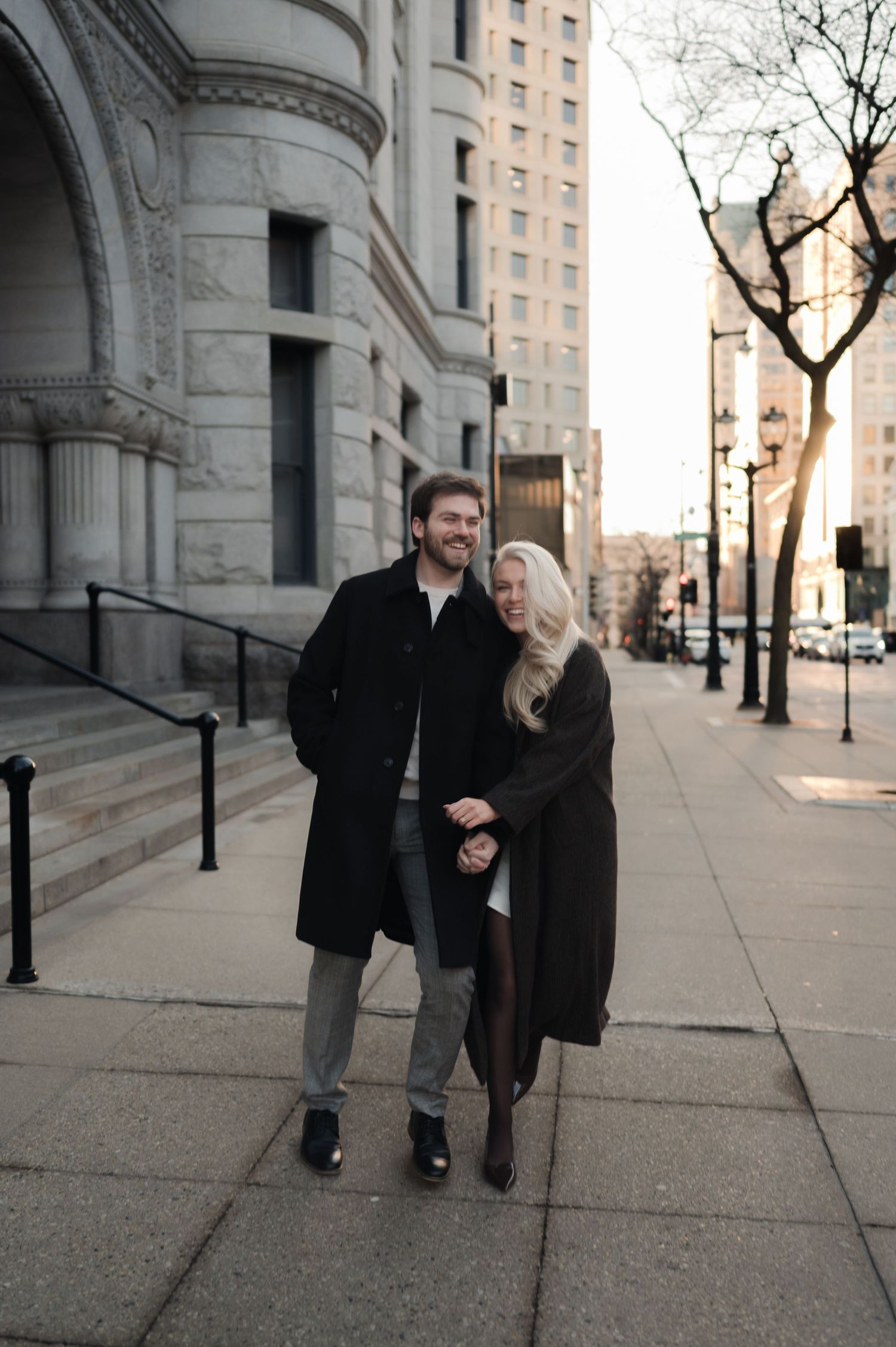 Happy couple walking hand in hand on a city sidewalk near a historic stone building at sunset.