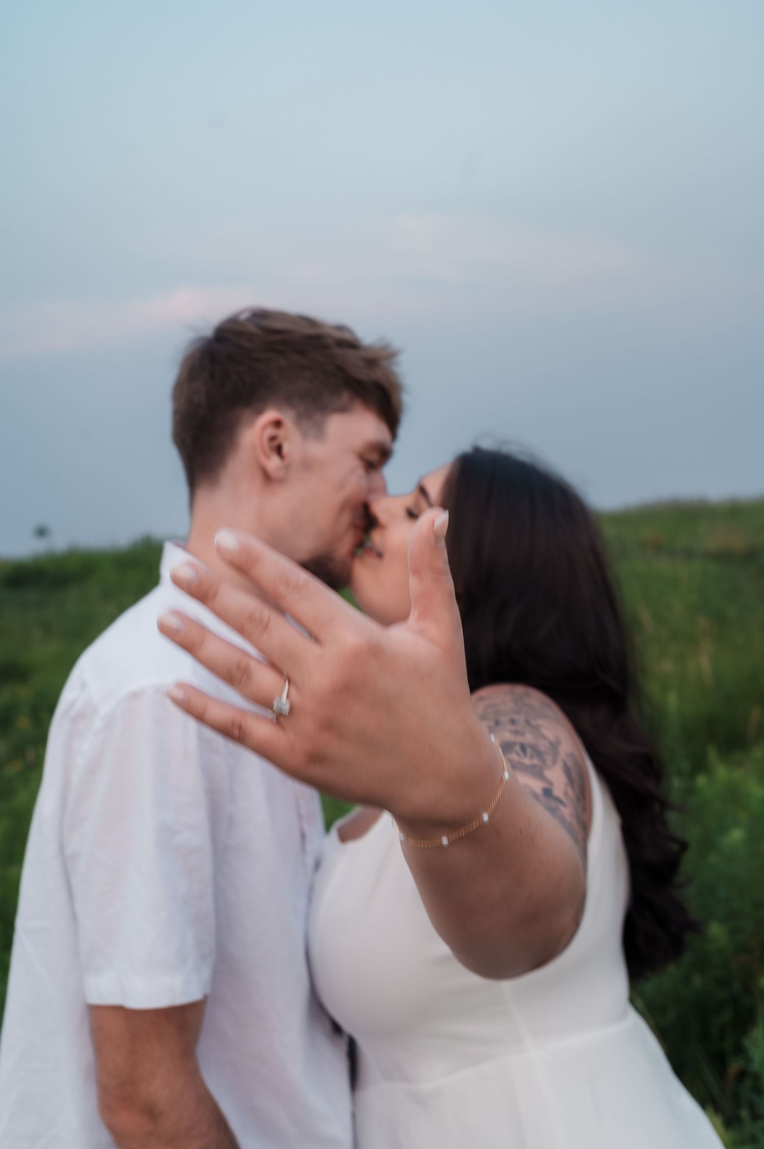 Couple kissing in a field while woman displays engagement ring on outstretched hand.