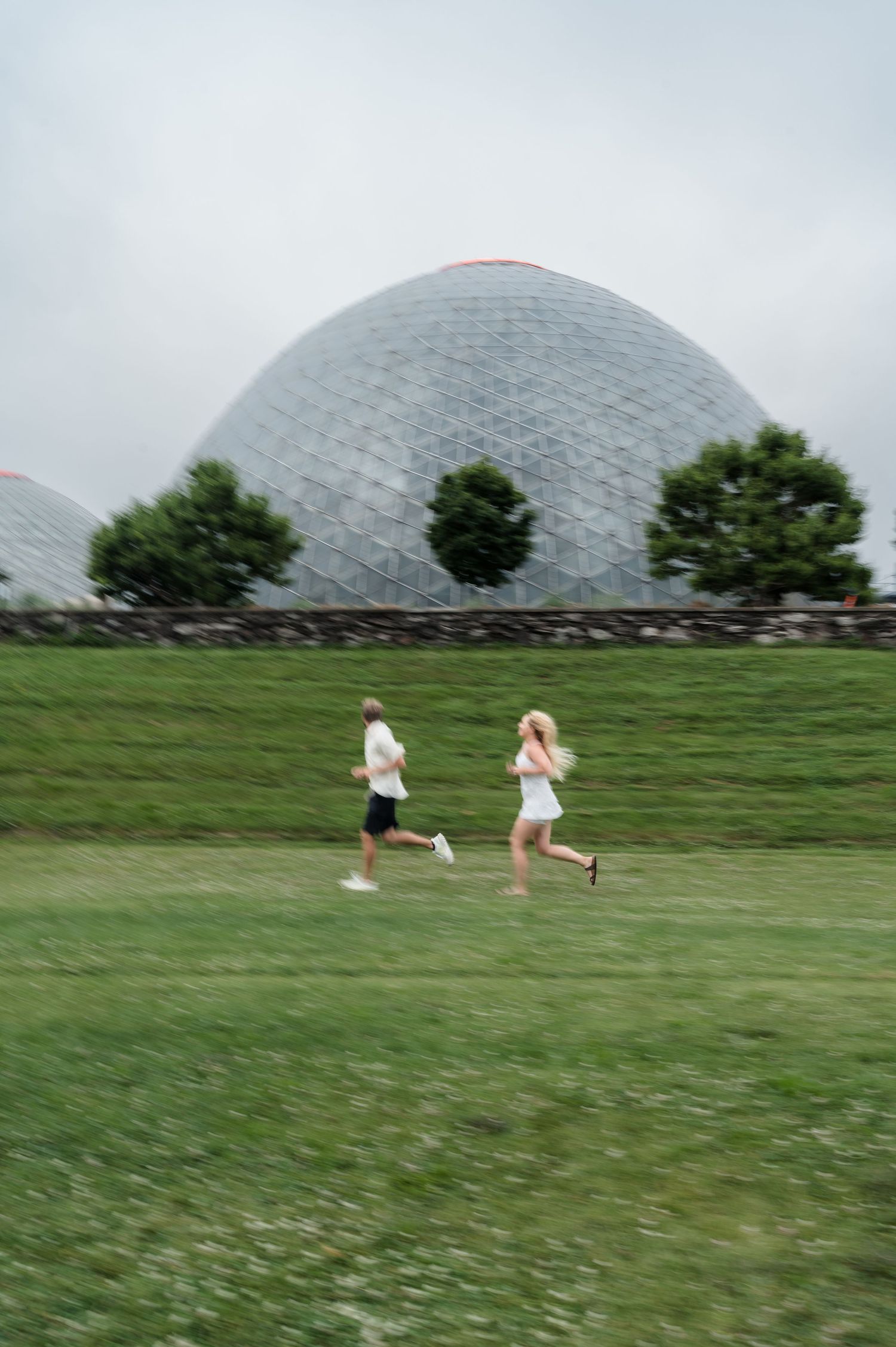 Couple running playfully across green grass with a large geodesic dome in the background.