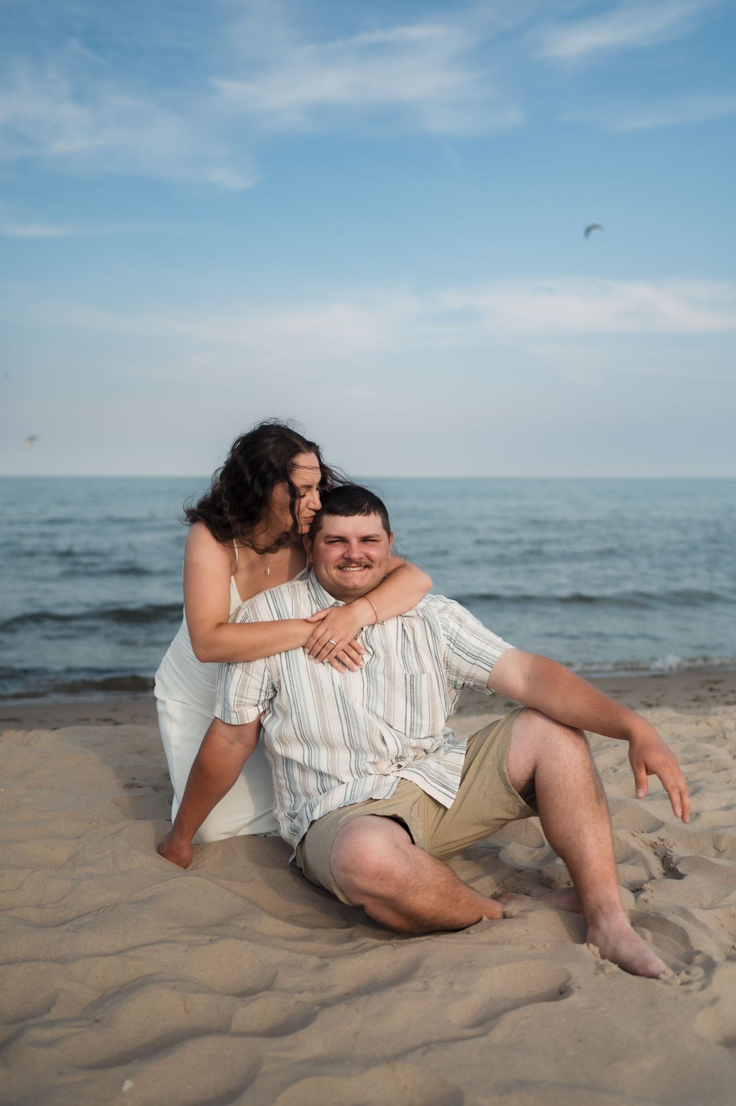 A couple shares a romantic moment sitting together on a sandy beach with the ocean and blue sky in the background.