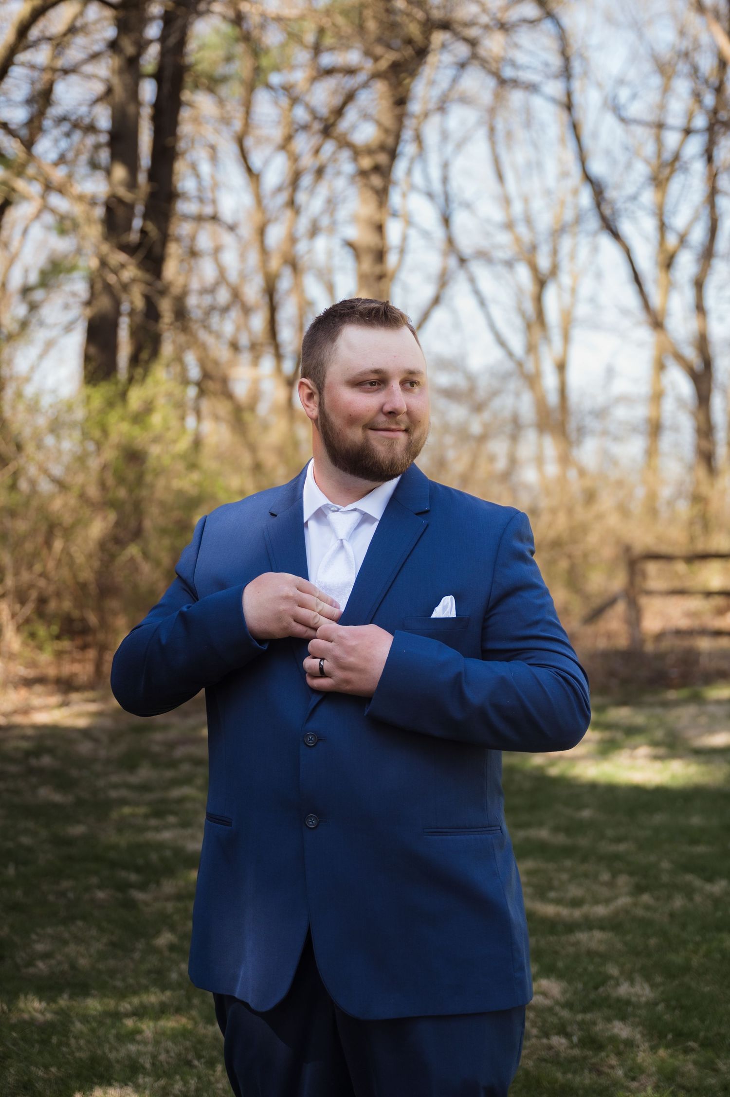 A person in a navy blue suit and white shirt stands outdoors adjusting their jacket in a wooded setting.