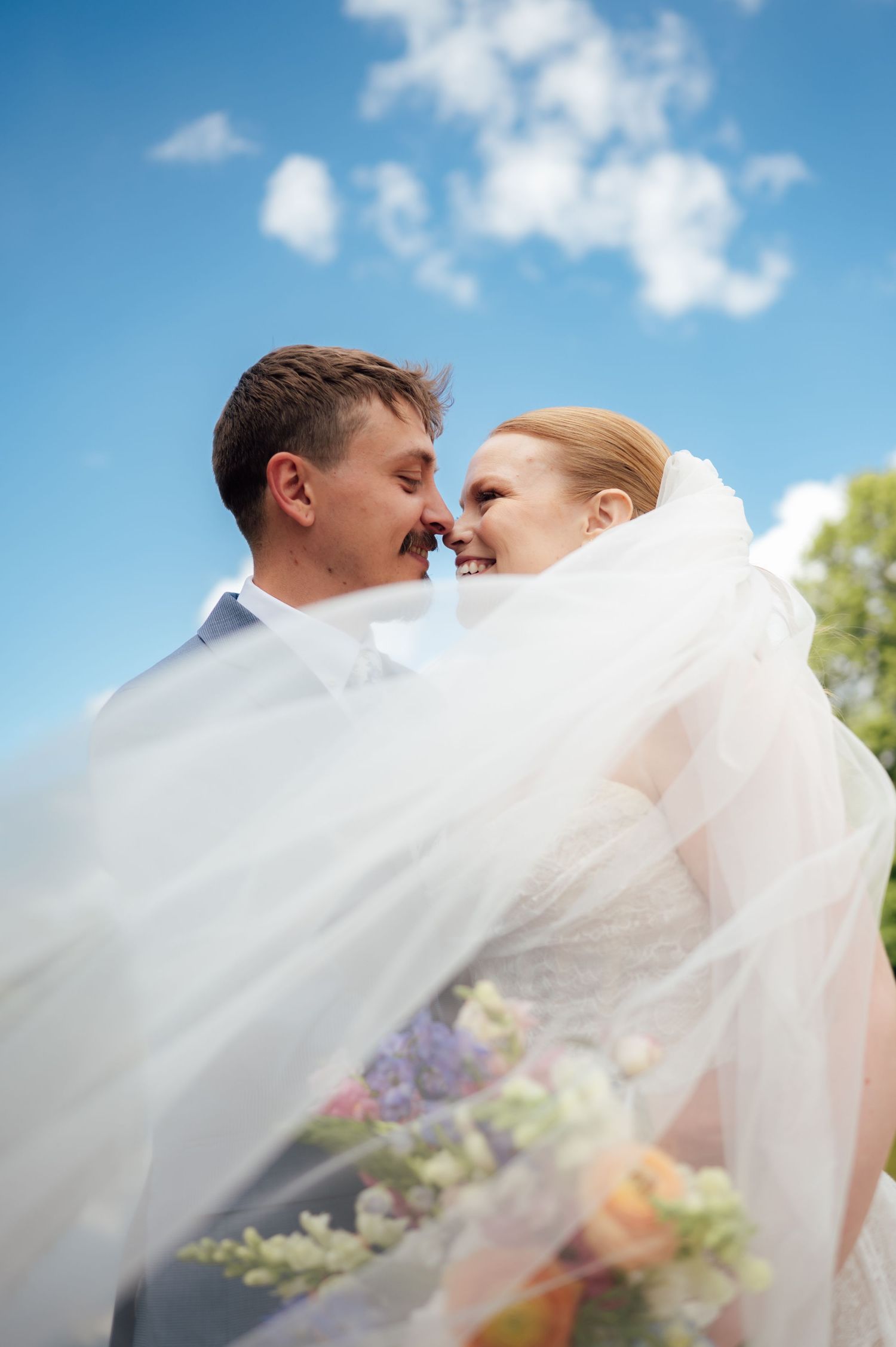 Bride and groom share a romantic moment under flowing white veil with blue sky backdrop on their wedding day.