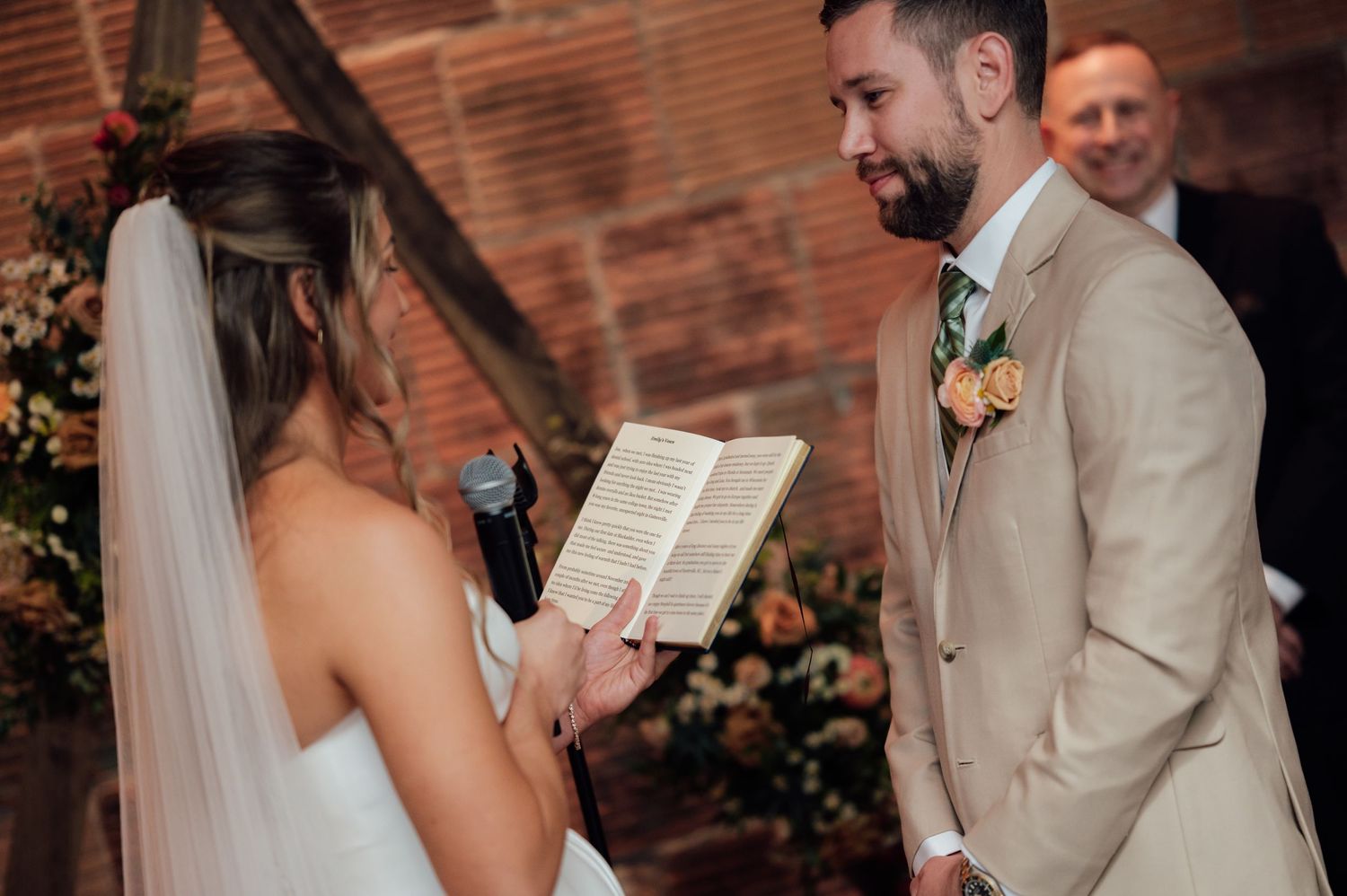 Bride reading wedding vows from a book to her smiling groom in a tan suit during an indoor ceremony.