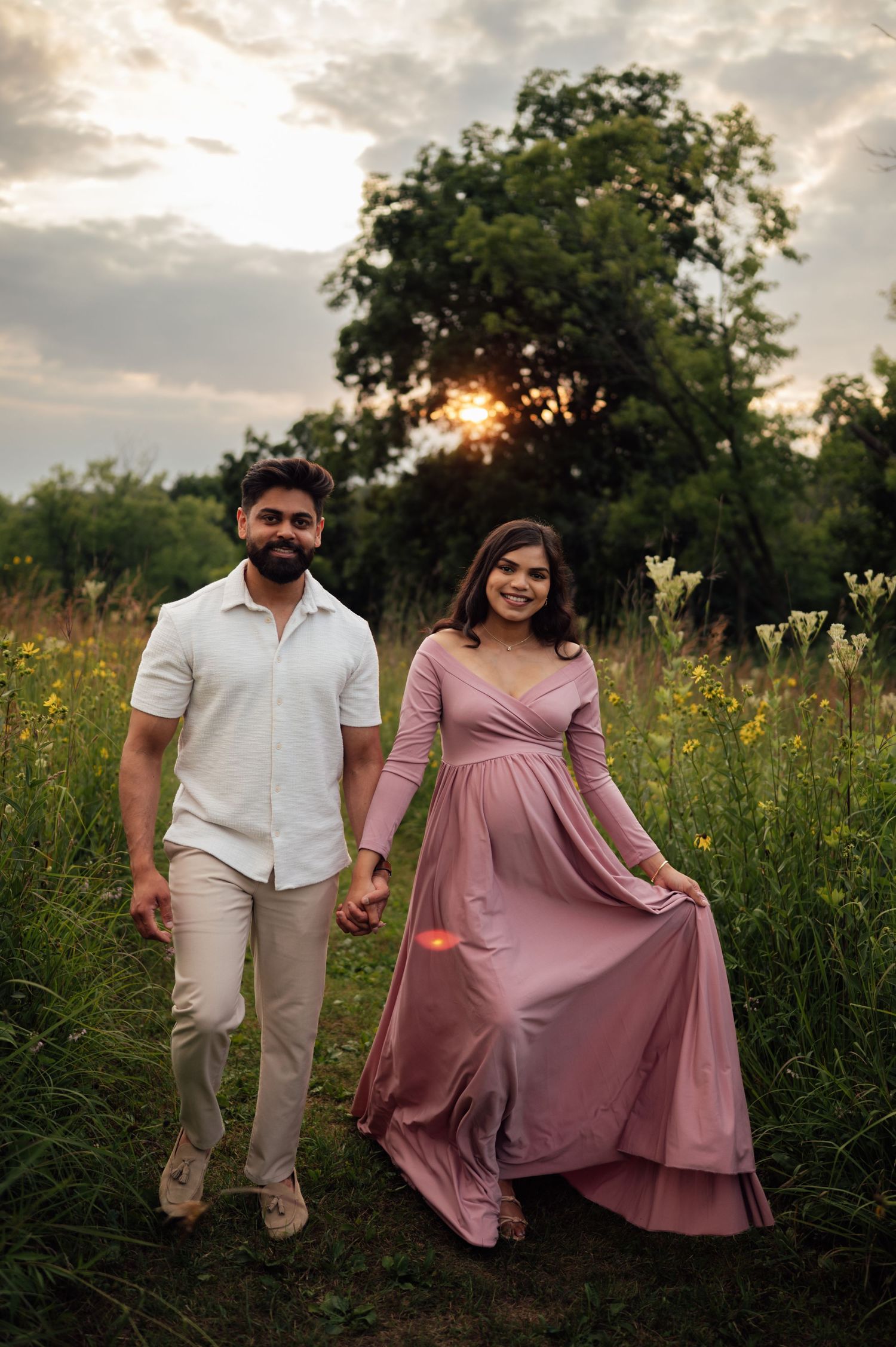 Expecting couple holds hands walking through wildflower meadow at golden sunset, woman wearing flowing pink maternity dress.