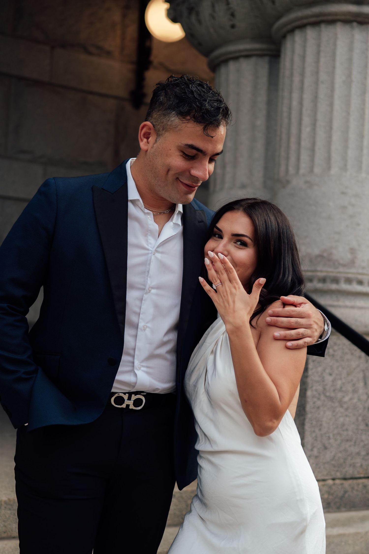 Couple celebrating engagement on stone steps, woman in white dress showing ring, man in navy suit smiling.