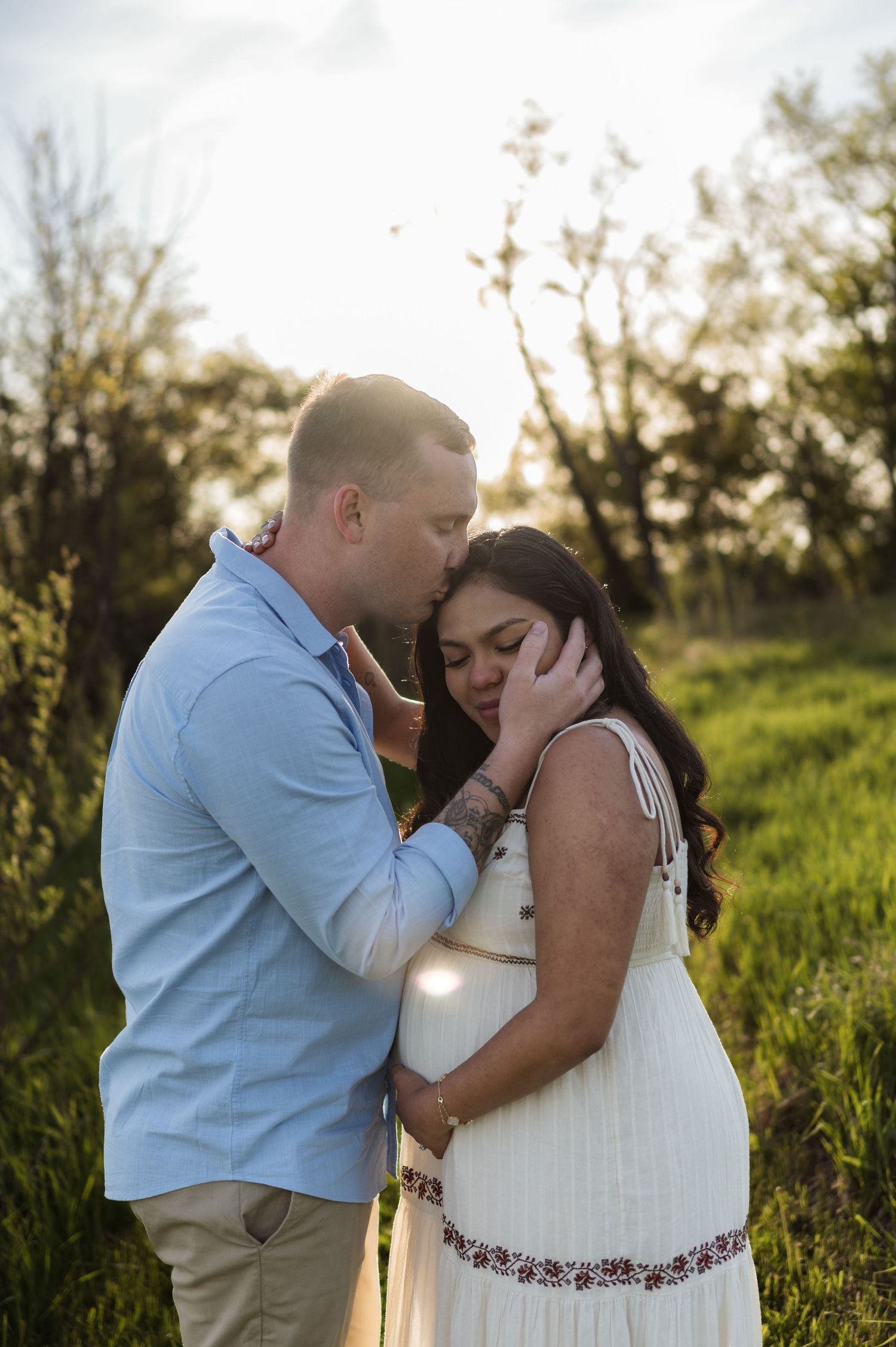 Man tenderly kissing pregnant woman's forehead while cradling her bump in sun-drenched outdoor field.