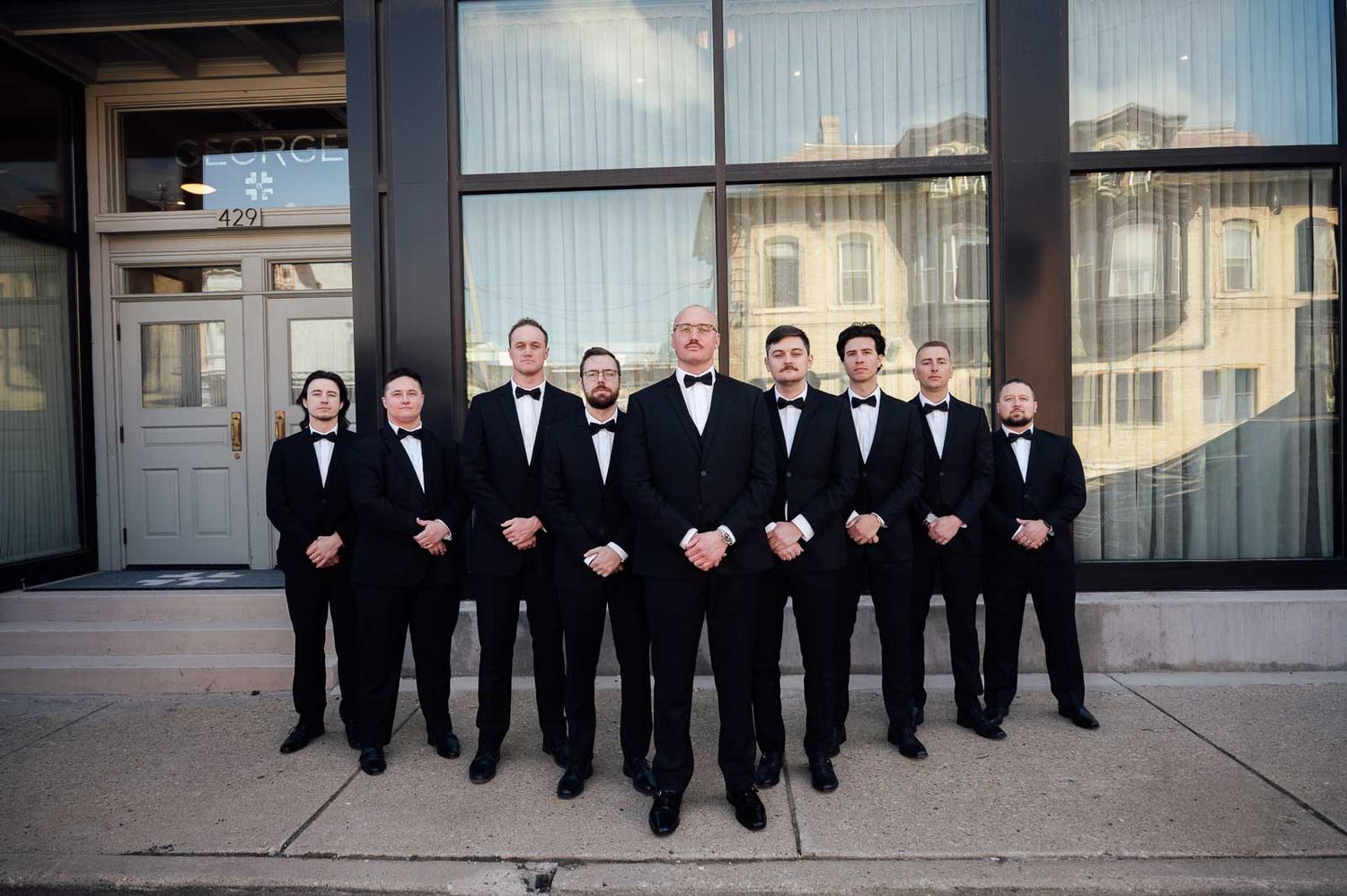 Groom and groomsmen posing in matching black tuxedos and bow ties outside urban wedding venue.