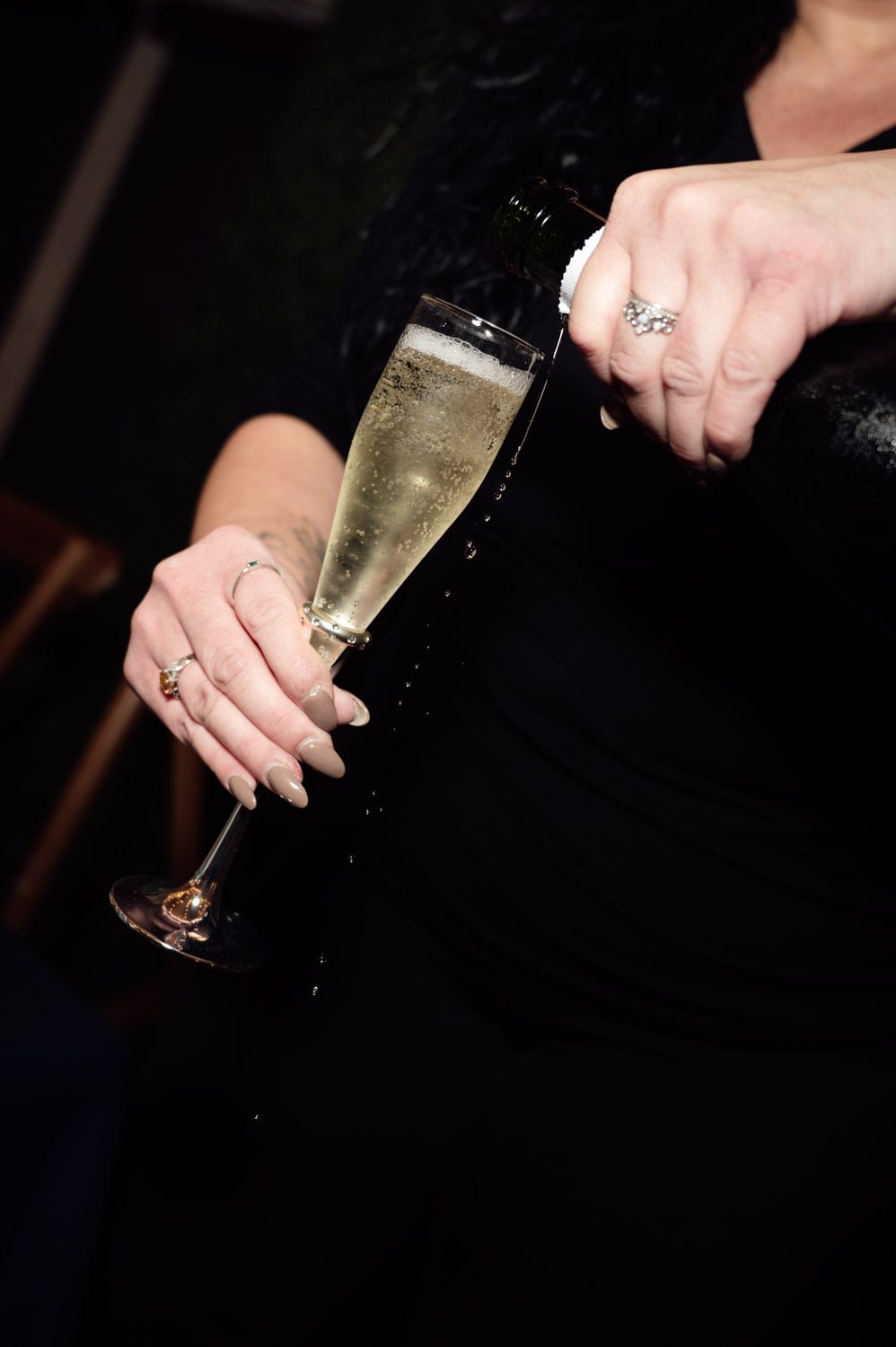 Woman in black dress pouring champagne into elegant flute glass wearing diamond engagement ring.