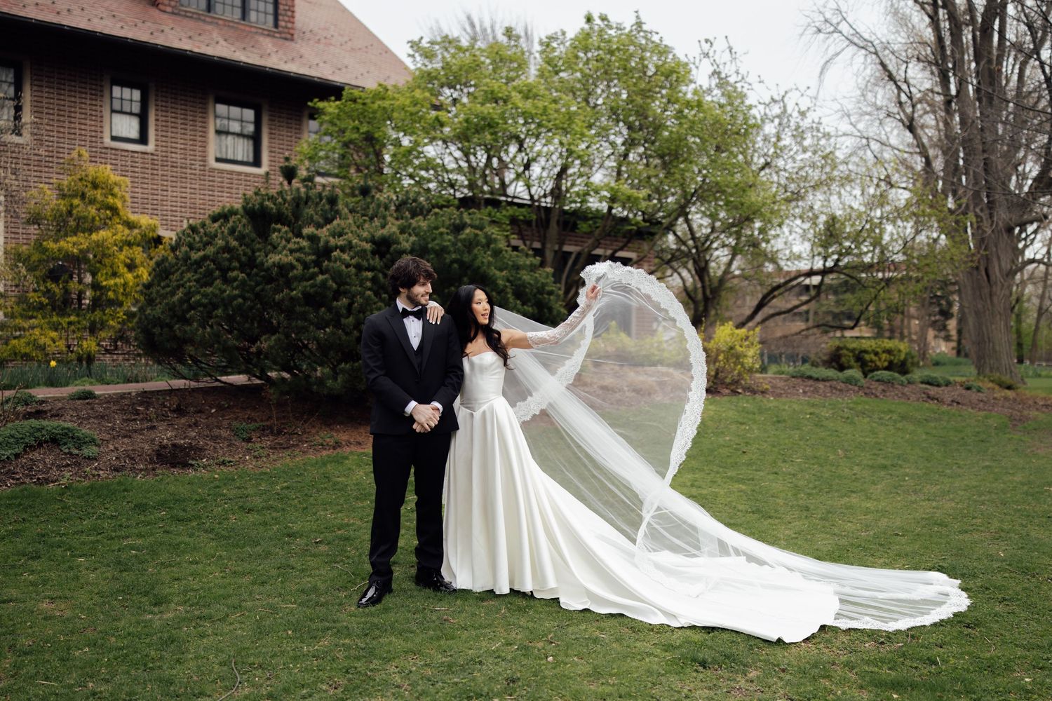 Bride and groom pose outdoors as cathedral veil billows dramatically in the breeze.