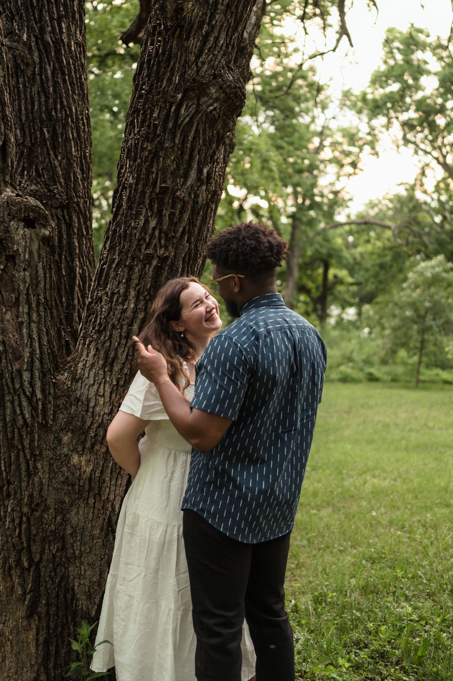 Interracial couple laughing together beside a large tree trunk in a lush green park setting.
