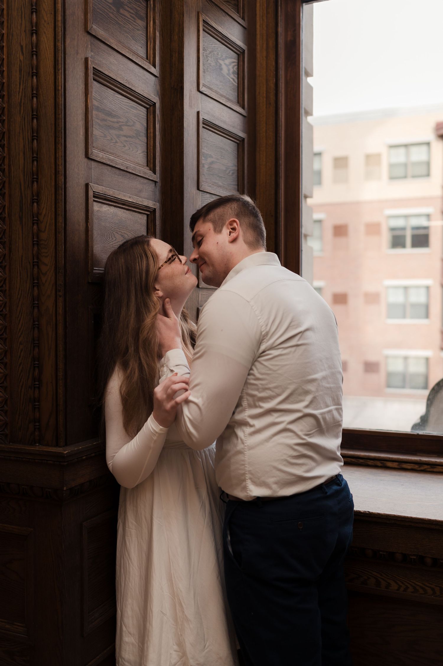 Couple sharing a tender kiss near a large wooden door and window in a warmly lit historic building.