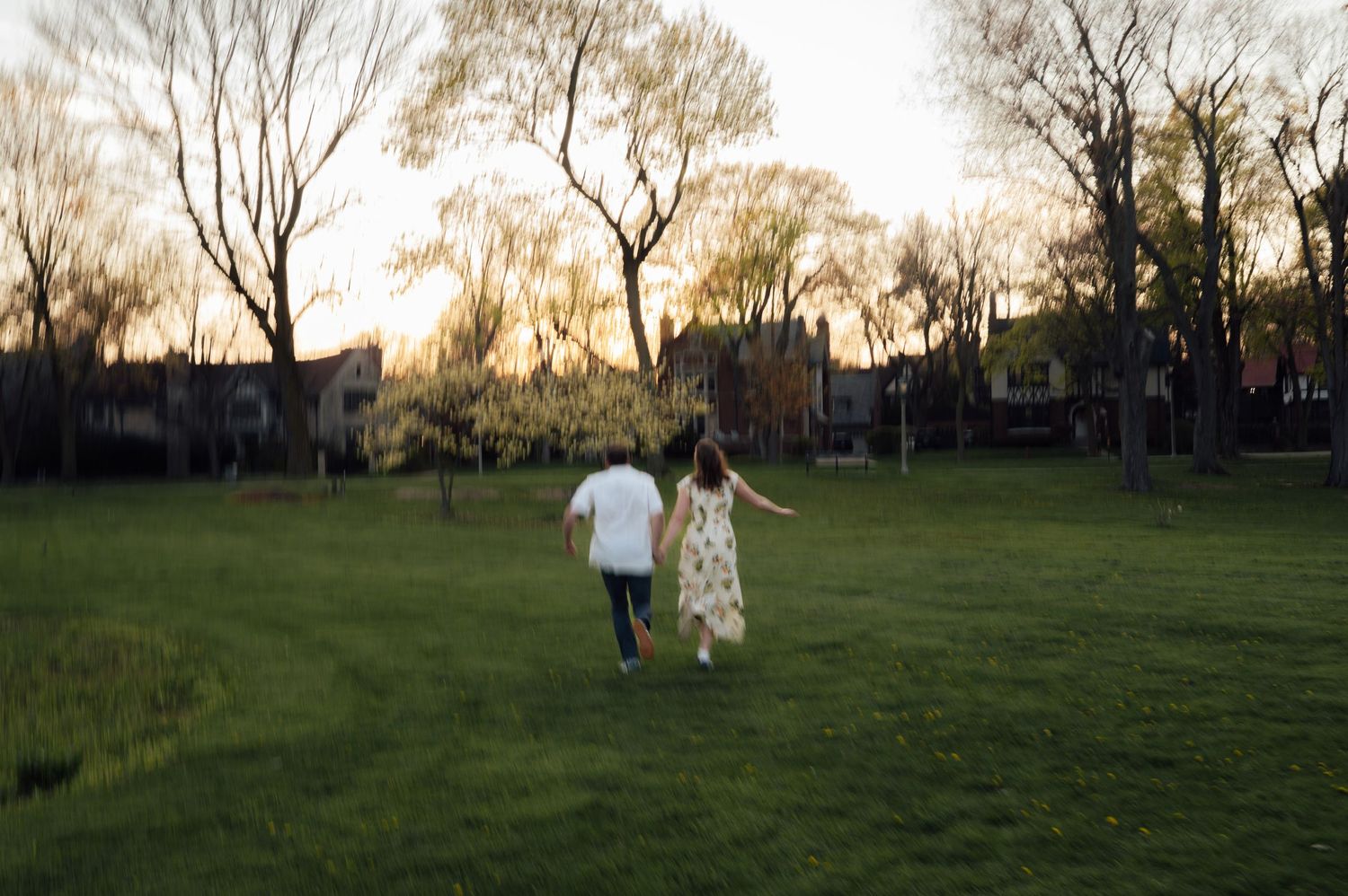 Couple holding hands running across a lush green park at golden hour with trees in the background.