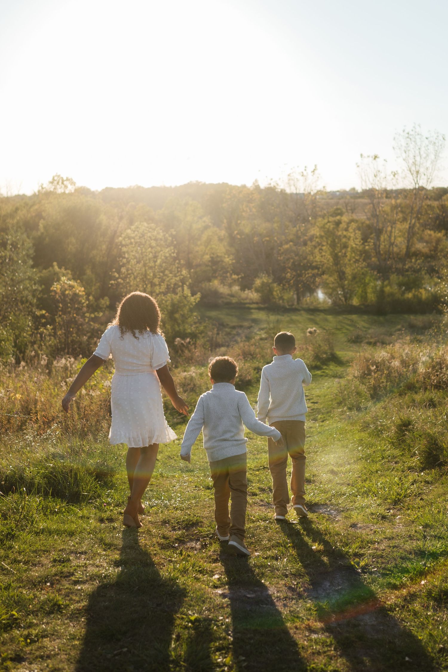 A mother in white holds hands with two boys walking toward a glowing sunset on a scenic outdoor path.
