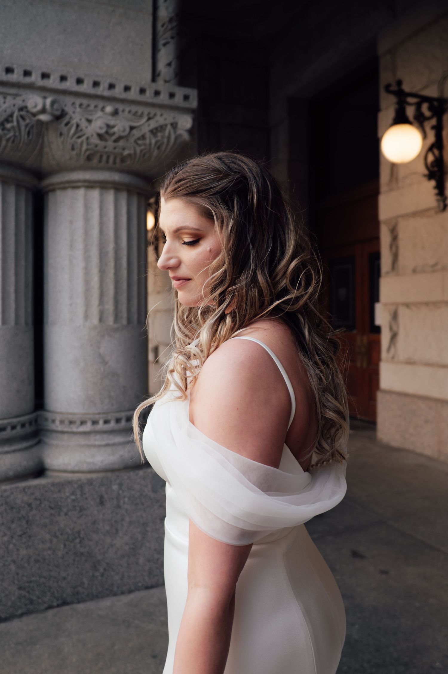 Bride in off-shoulder white gown with tulle sleeves poses near stone columns, hair in soft waves.