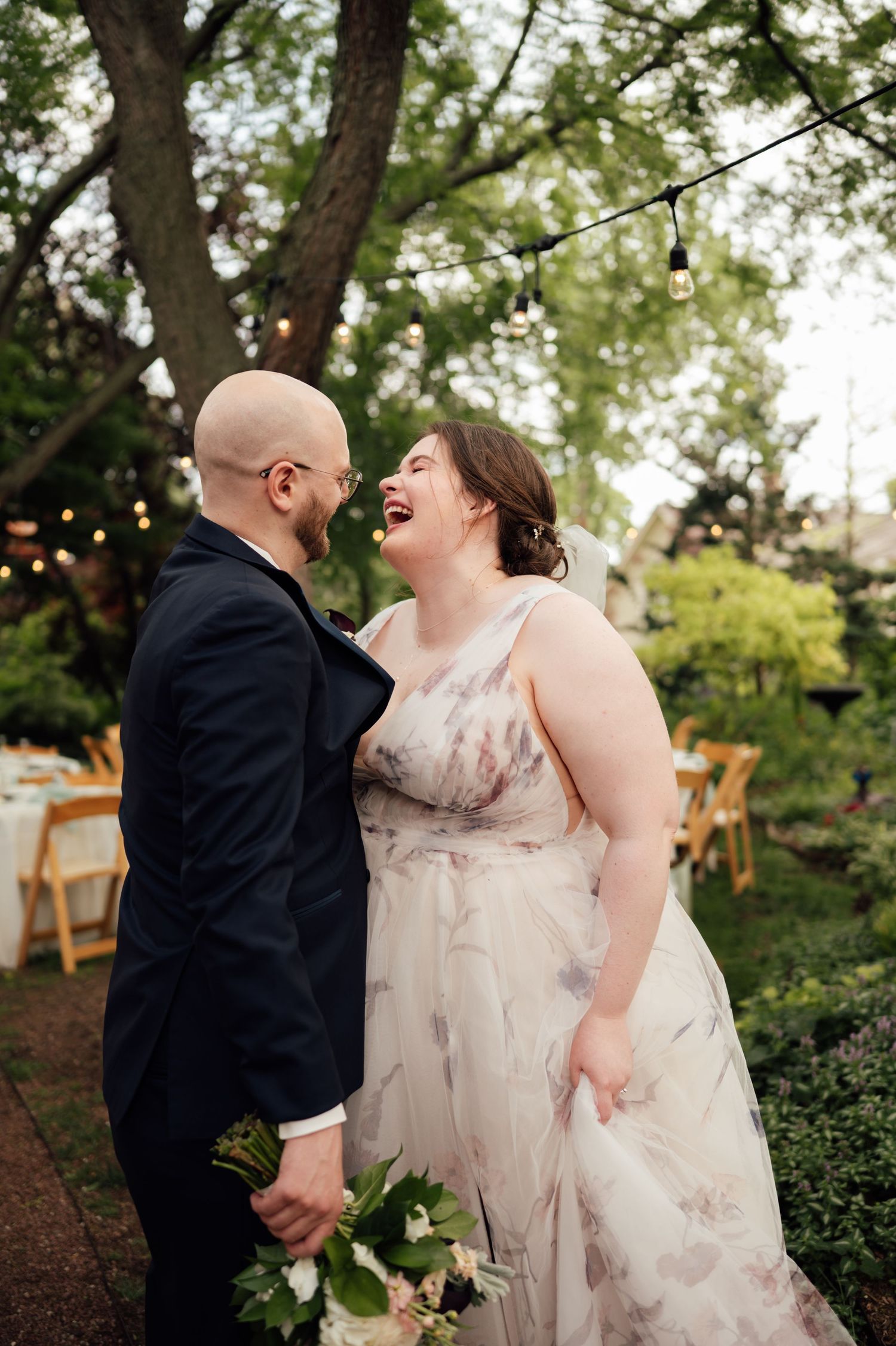 A joyful couple laughs together in a garden wedding setting, bride in floral gown holding bouquet, groom in navy suit.
