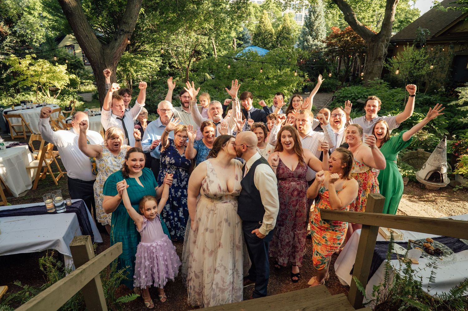 Joyful wedding group photo with guests cheering outdoors in a lush garden setting.