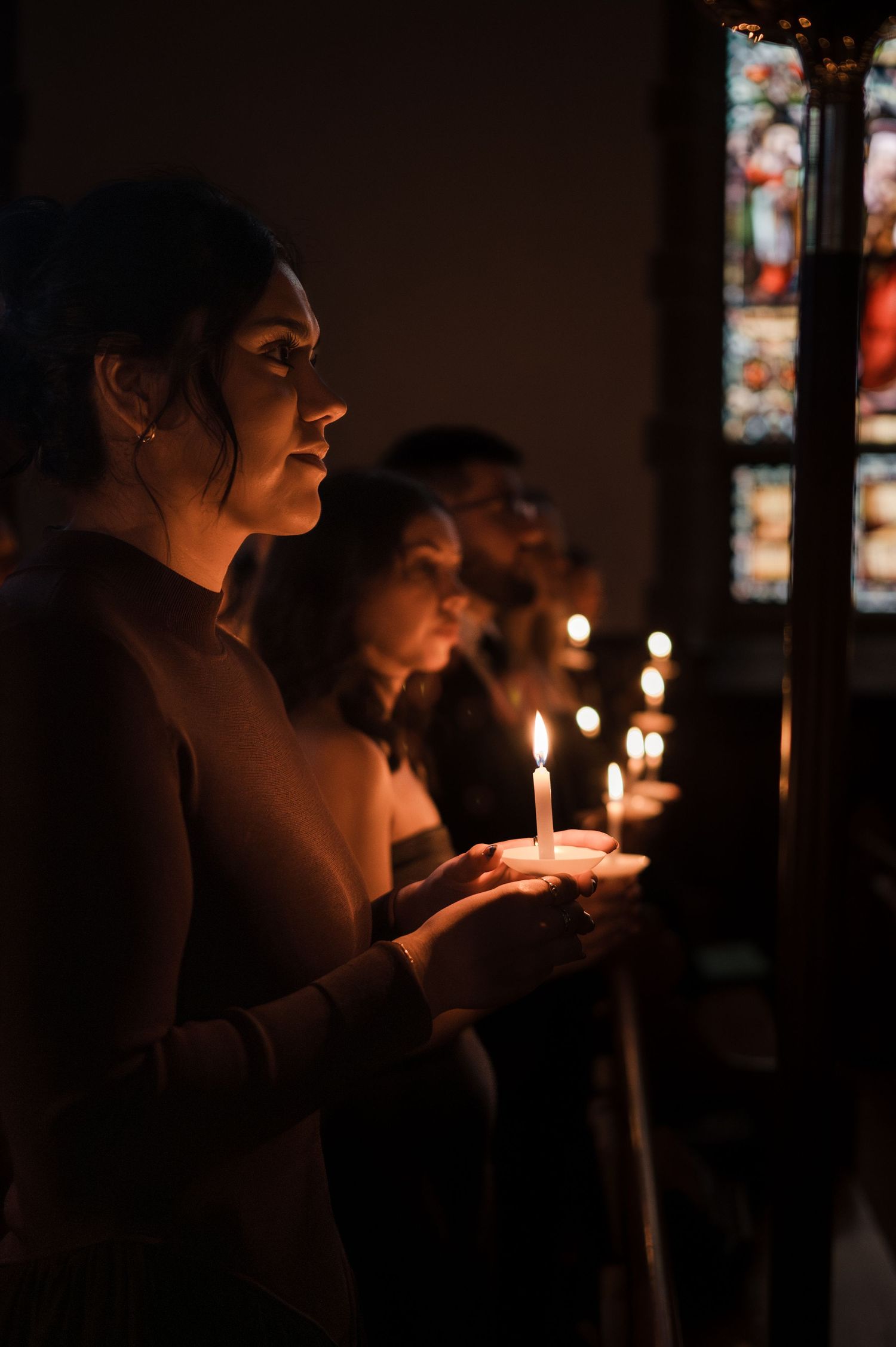 People holding lit candles during a candlelight church service, with stained glass windows glowing in the background.