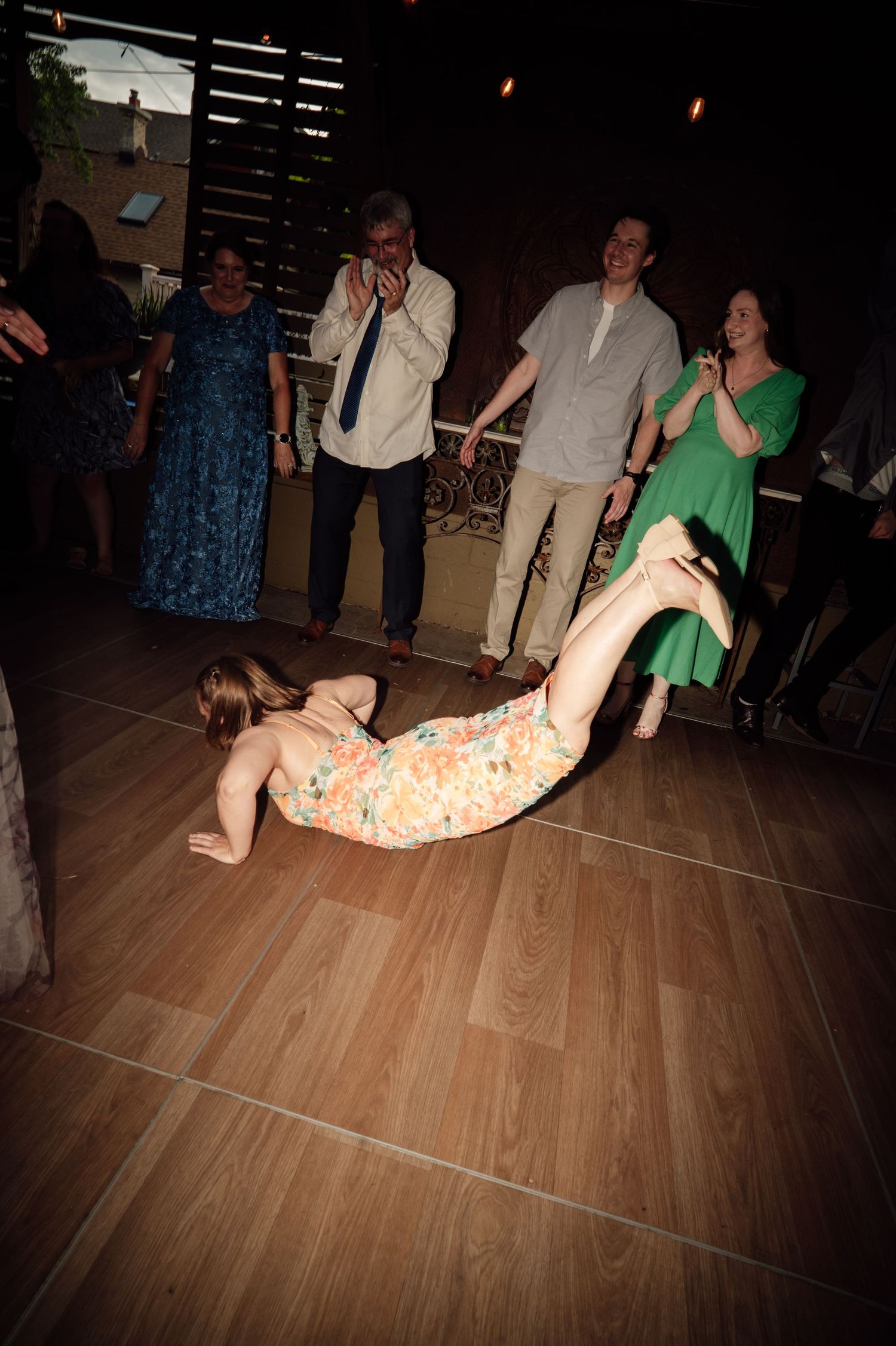 A woman in a floral dress falls on a dance floor while wedding guests laugh and react with surprise behind her.