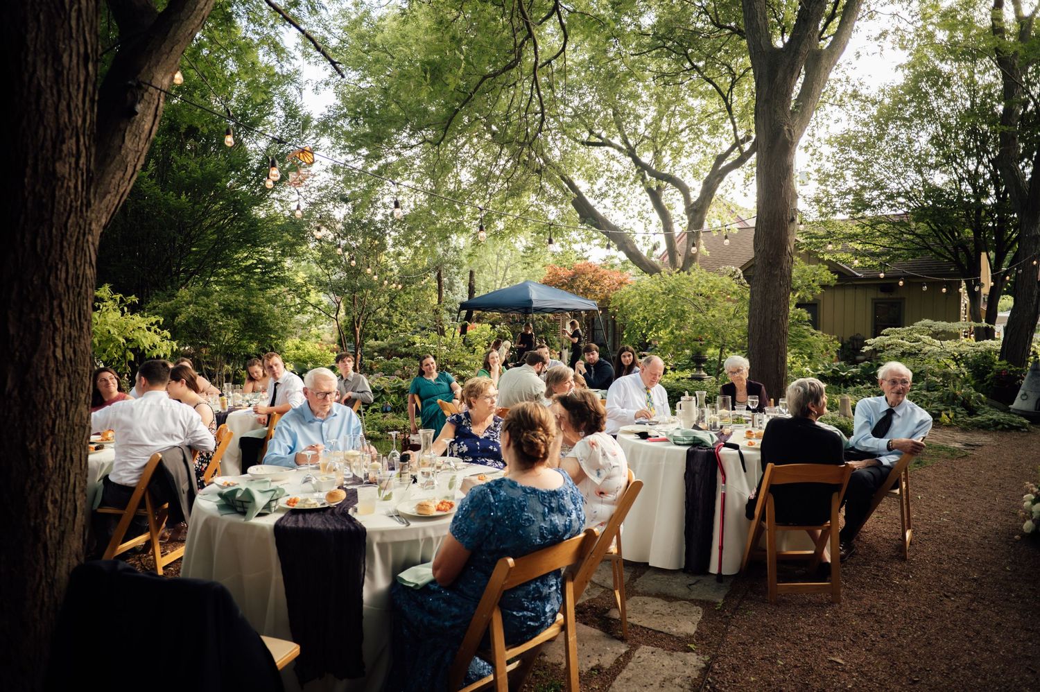 Guests dining outdoors at elegant wedding reception tables surrounded by lush trees in a garden setting.