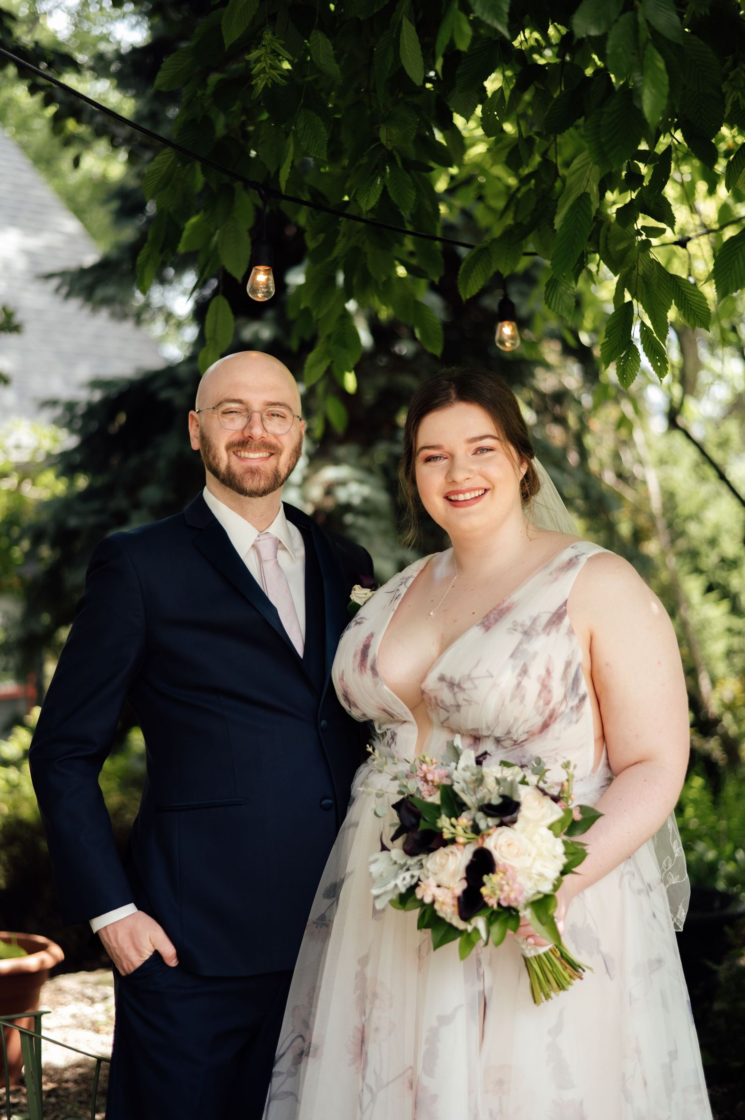Bride and groom smiling outdoors under string lights, bride holding white and purple bouquet in floral wedding gown.