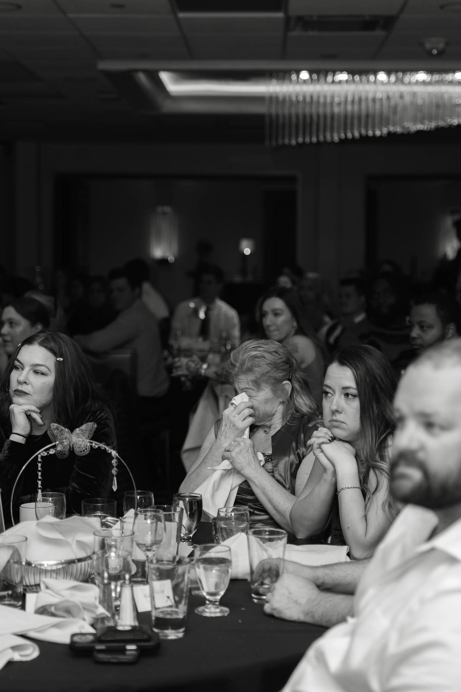 Emotional wedding guests wipe tears during heartfelt speeches at an elegant ballroom reception dinner.