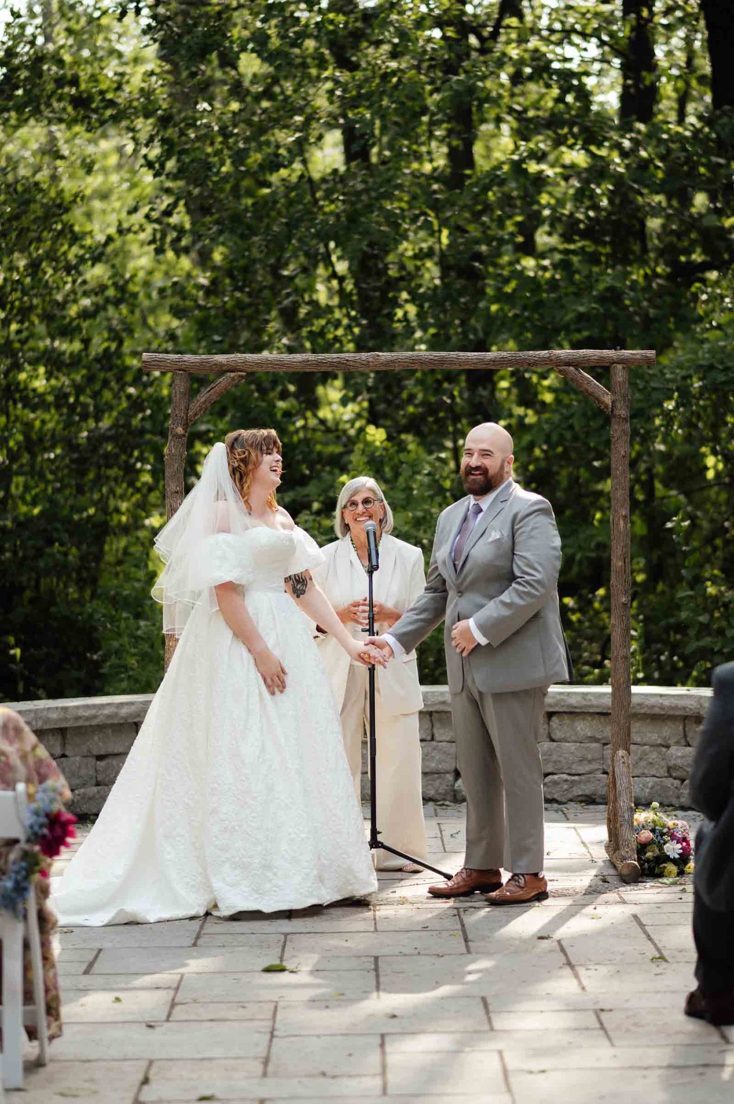 Bride and groom exchanging vows under rustic wooden arch at outdoor ceremony.