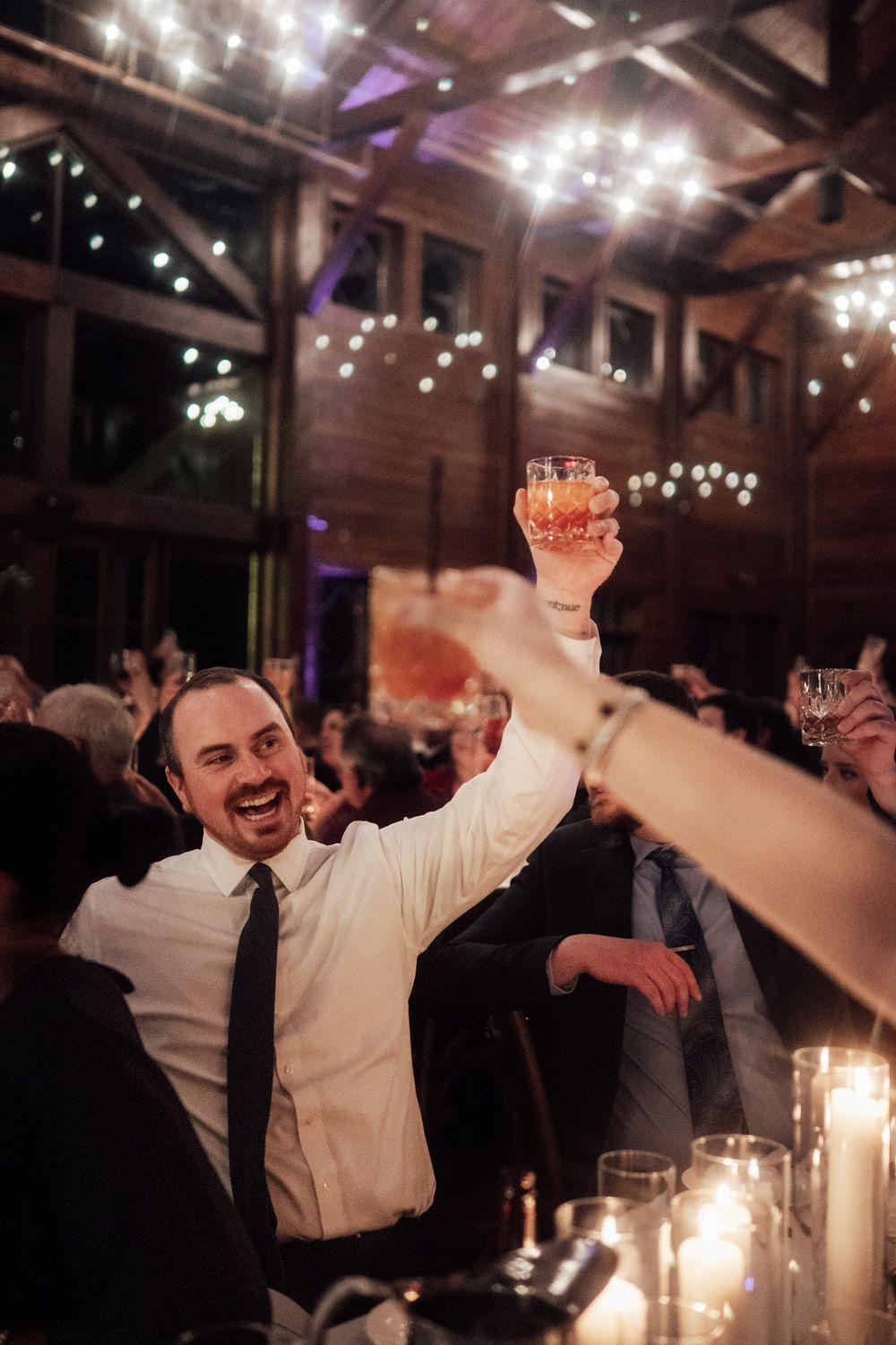 Smiling wedding guest raising whiskey glass in toast at candlelit rustic barn reception.